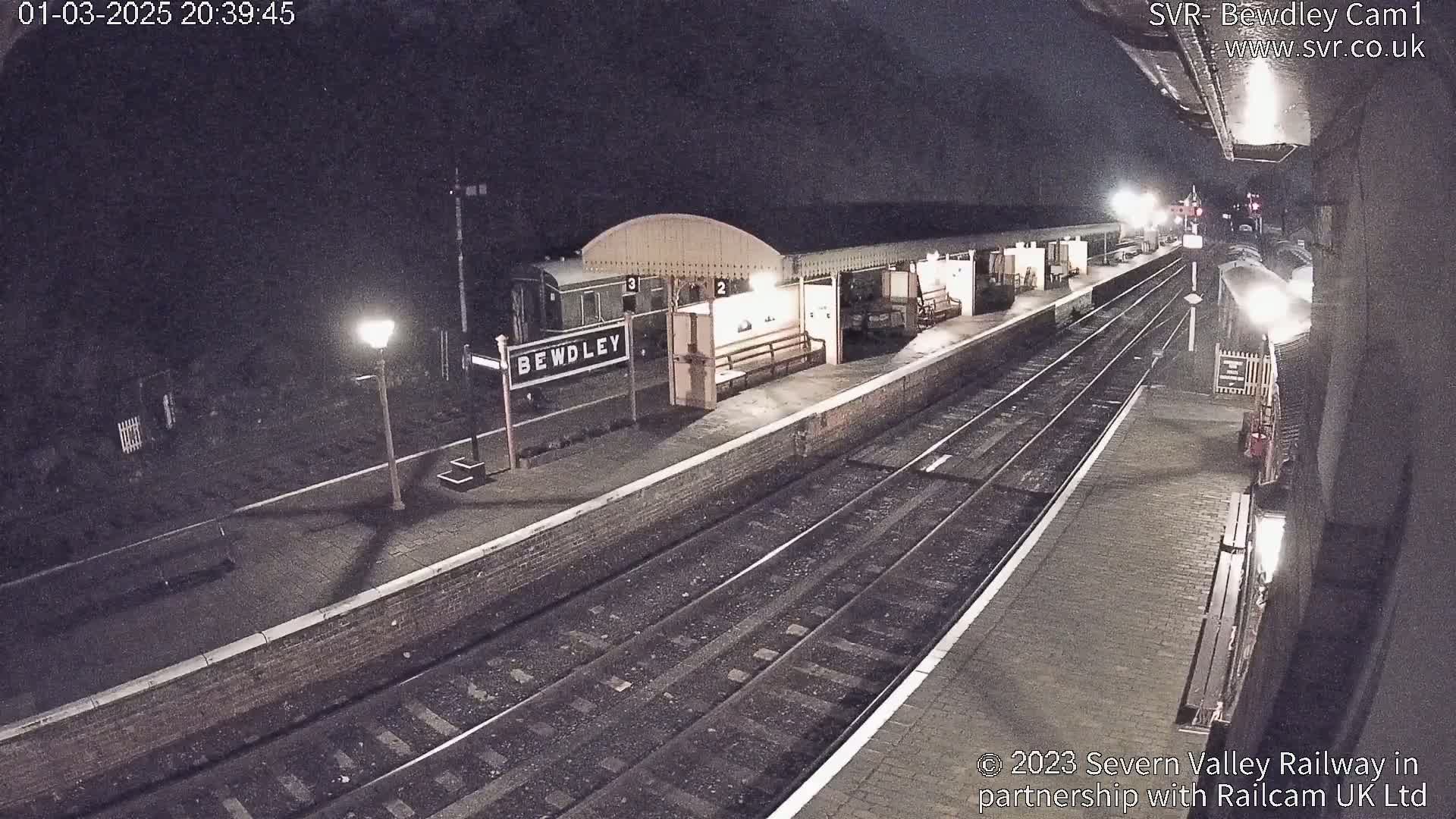 A nighttime view of a train station platform with railway tracks, a train partially visible, and a lit lamppost.