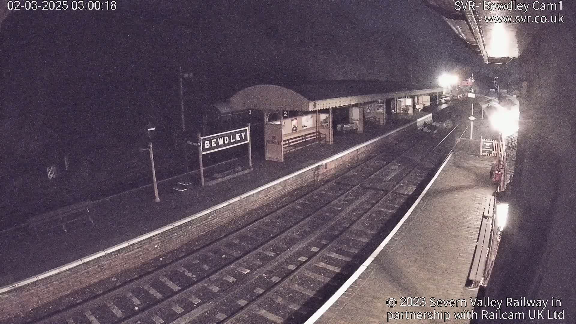 A nighttime view of a train station platform with a train arriving, under dark skies.