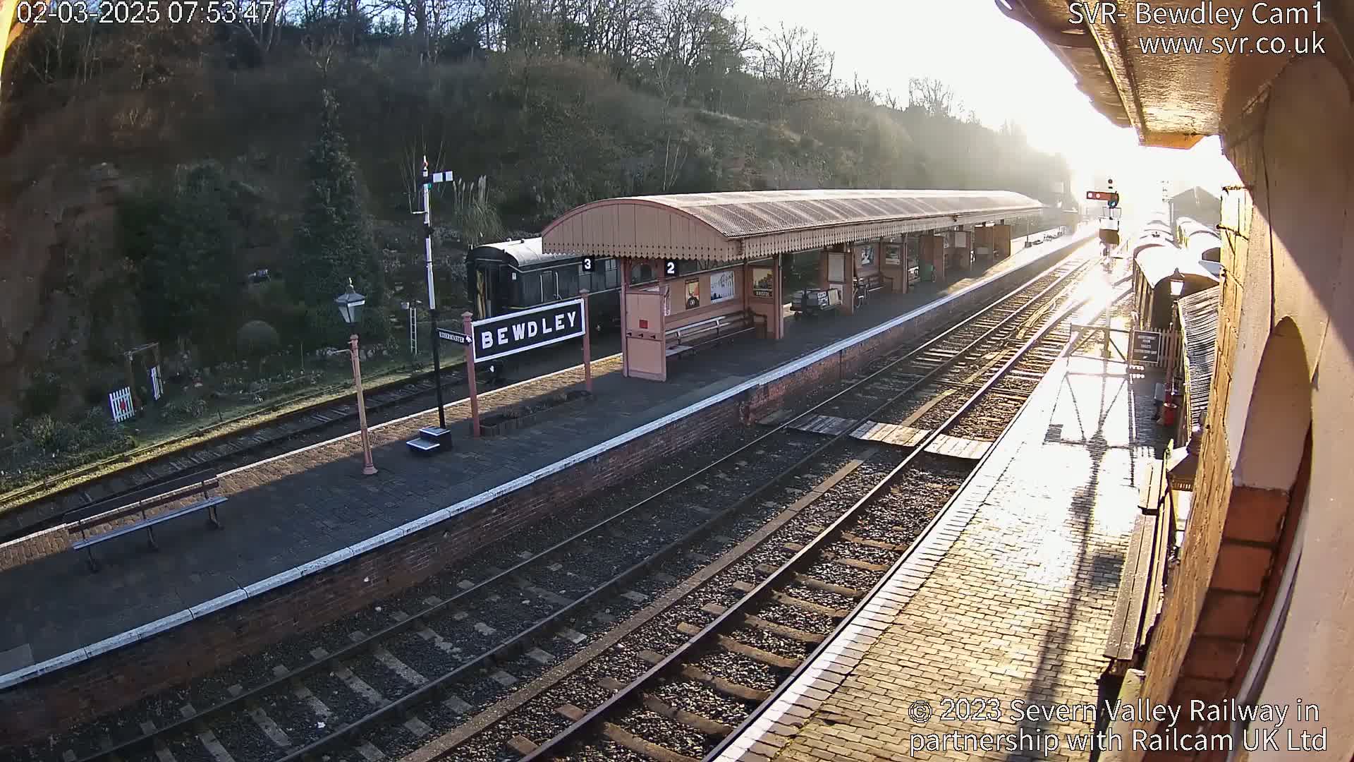 A sunny train station with a platform, railway tracks, and a train partially visible, sits next to a hillside with vegetation.