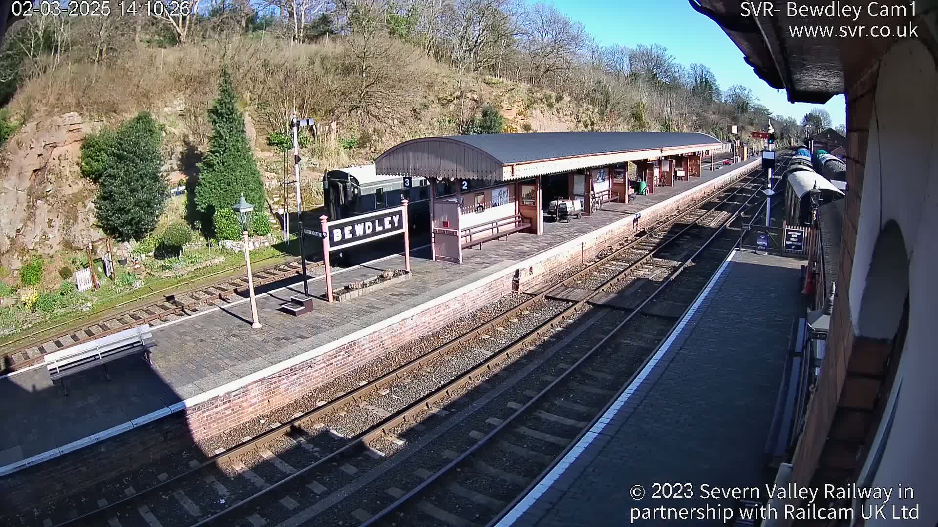 A train sits at a station platform on a sunny day, next to a brick building and several sets of train tracks.