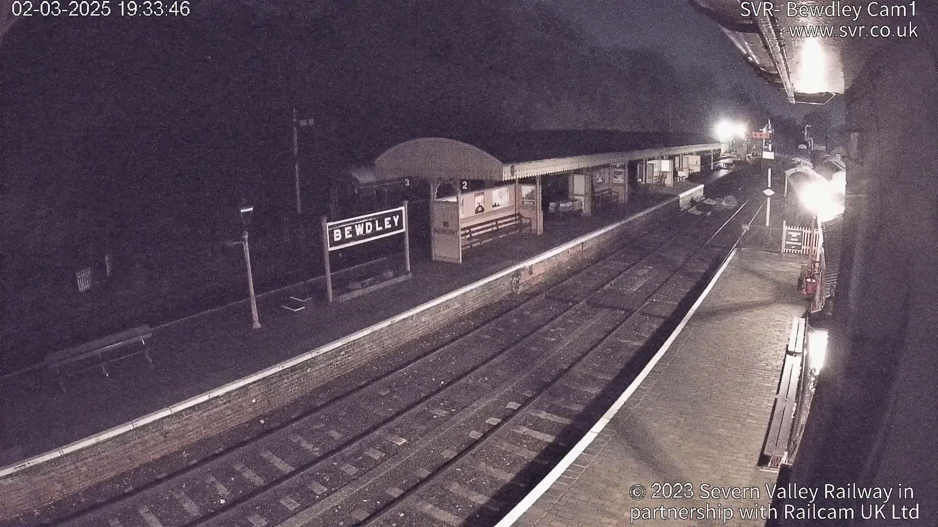 A nighttime view of a train station platform with train tracks and a partially visible train.