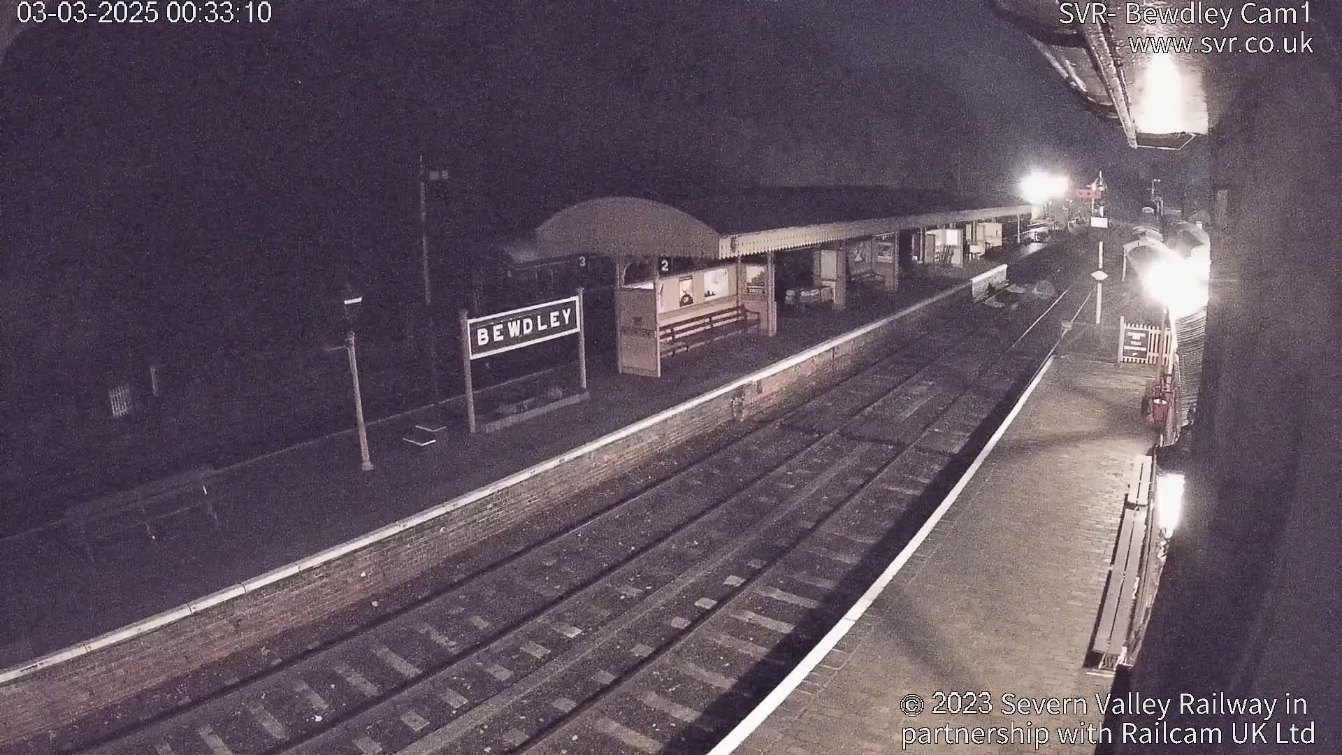 A nighttime view of a train station platform with a train partially visible, under dark conditions.