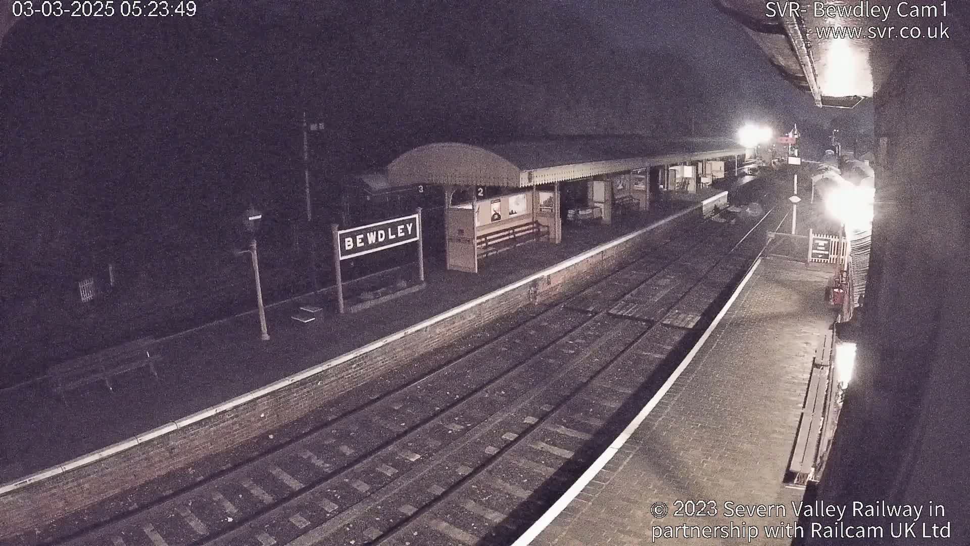 A nighttime view of a train station platform with train tracks and a waiting shelter under dark, possibly cloudy skies.
