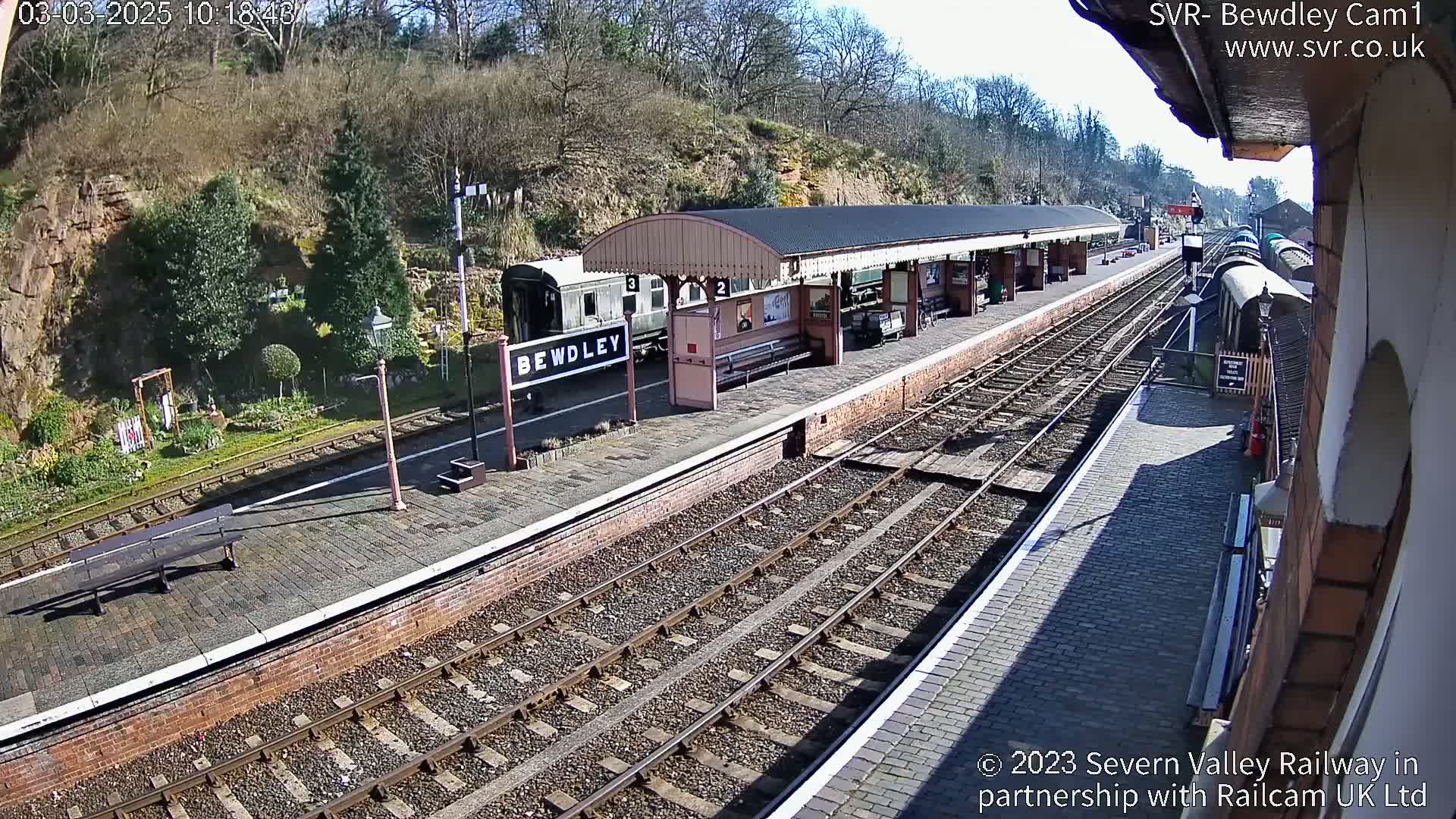 A train station platform with a covered waiting area, a train car, and multiple train tracks, all under a sunny sky.