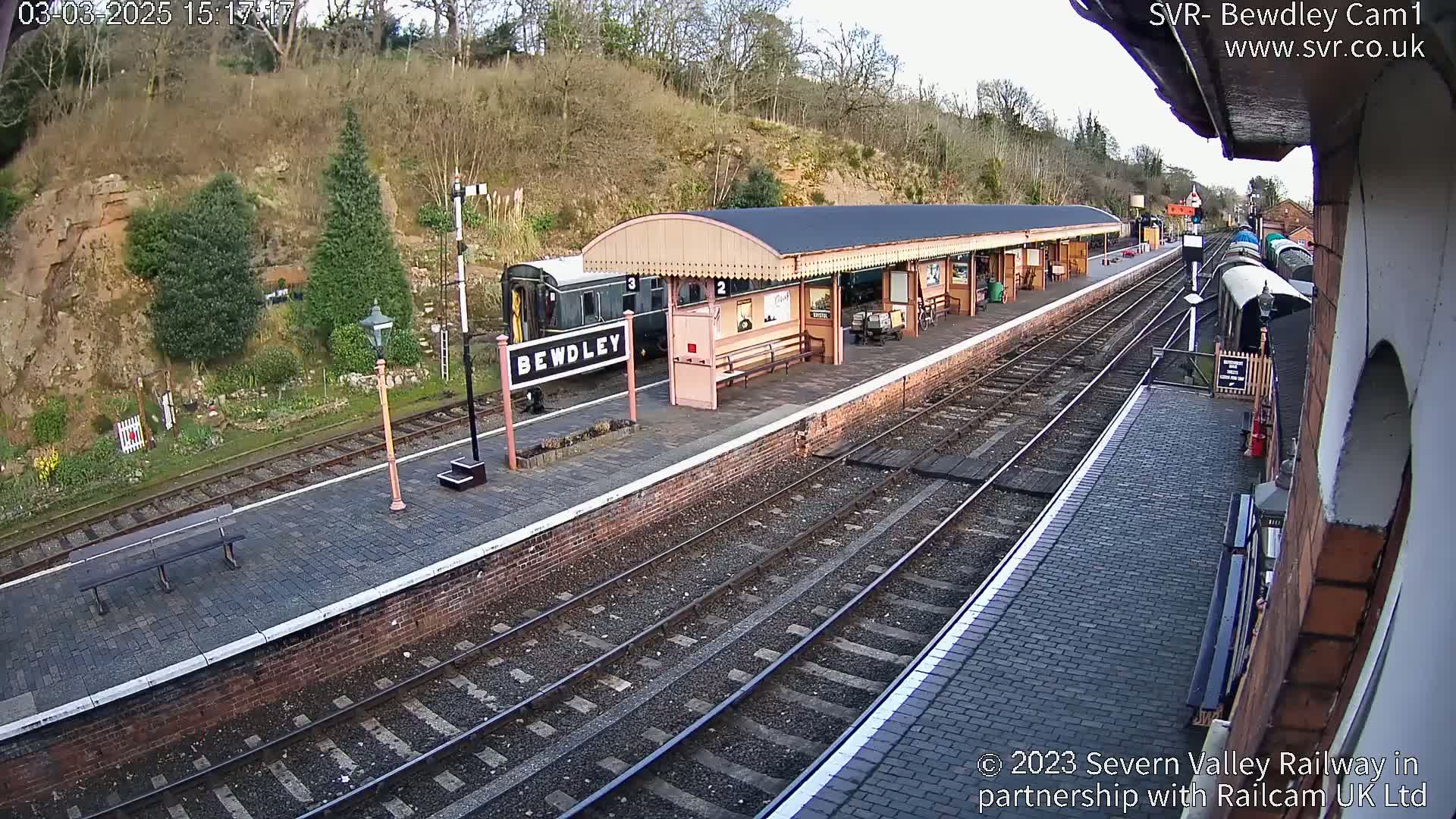 A train sits at a station platform next to a covered waiting area on a partly sunny day.