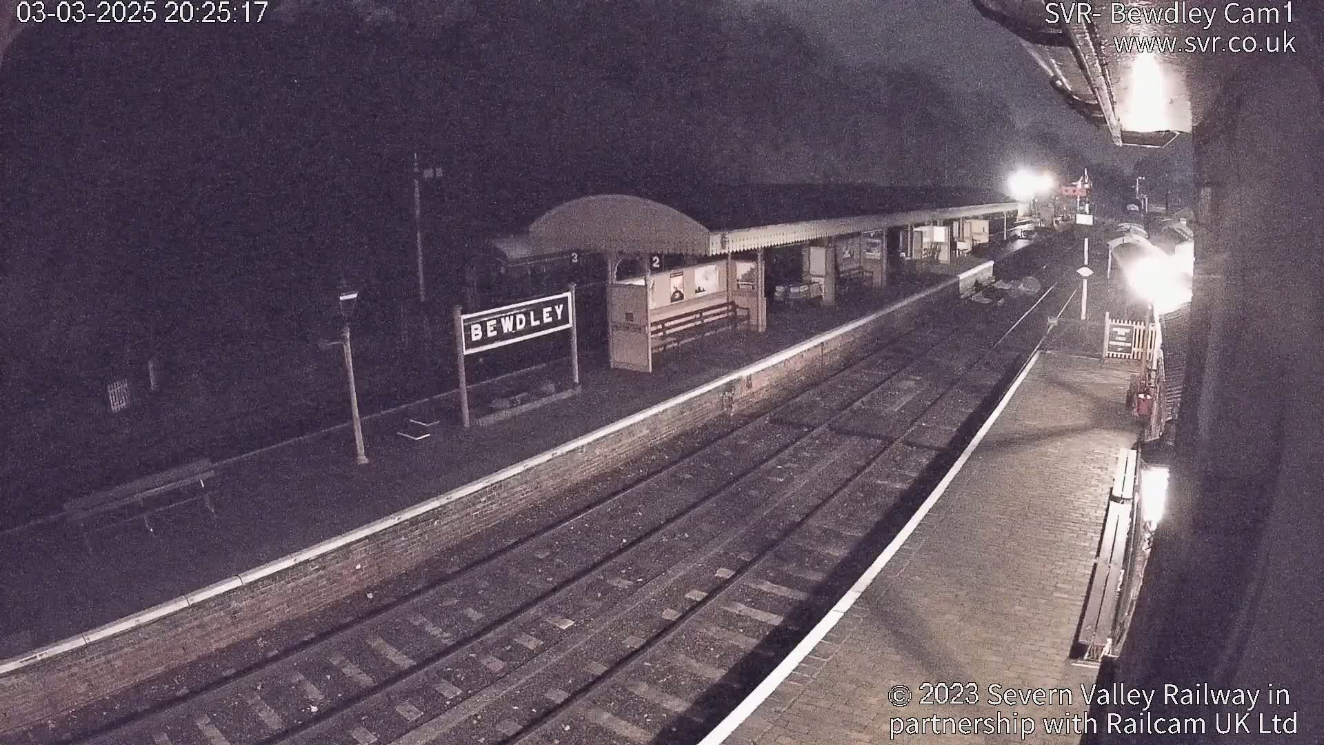 A nighttime view of a train station platform with train tracks and a partially visible train under a dark sky.