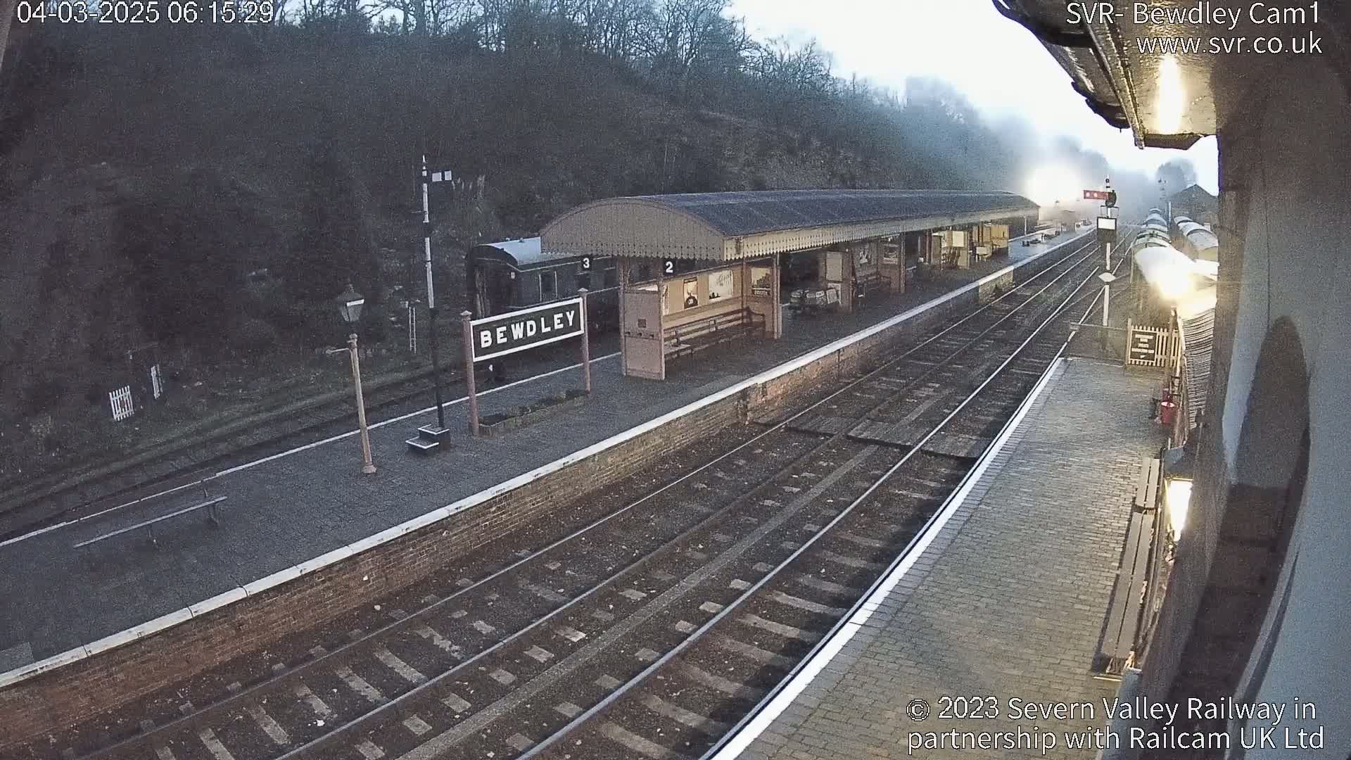 A steam train departs from a train station platform on a foggy morning.