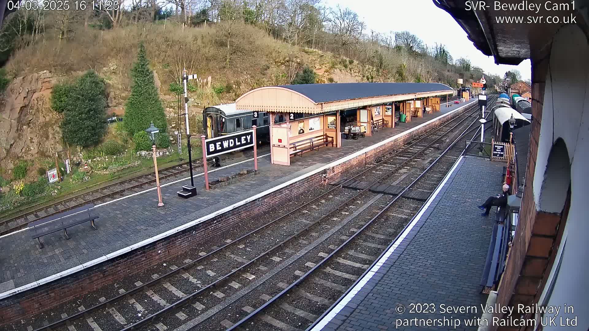 A train sits at a railway station platform on a partly sunny day, with a person sitting on a nearby wall.
