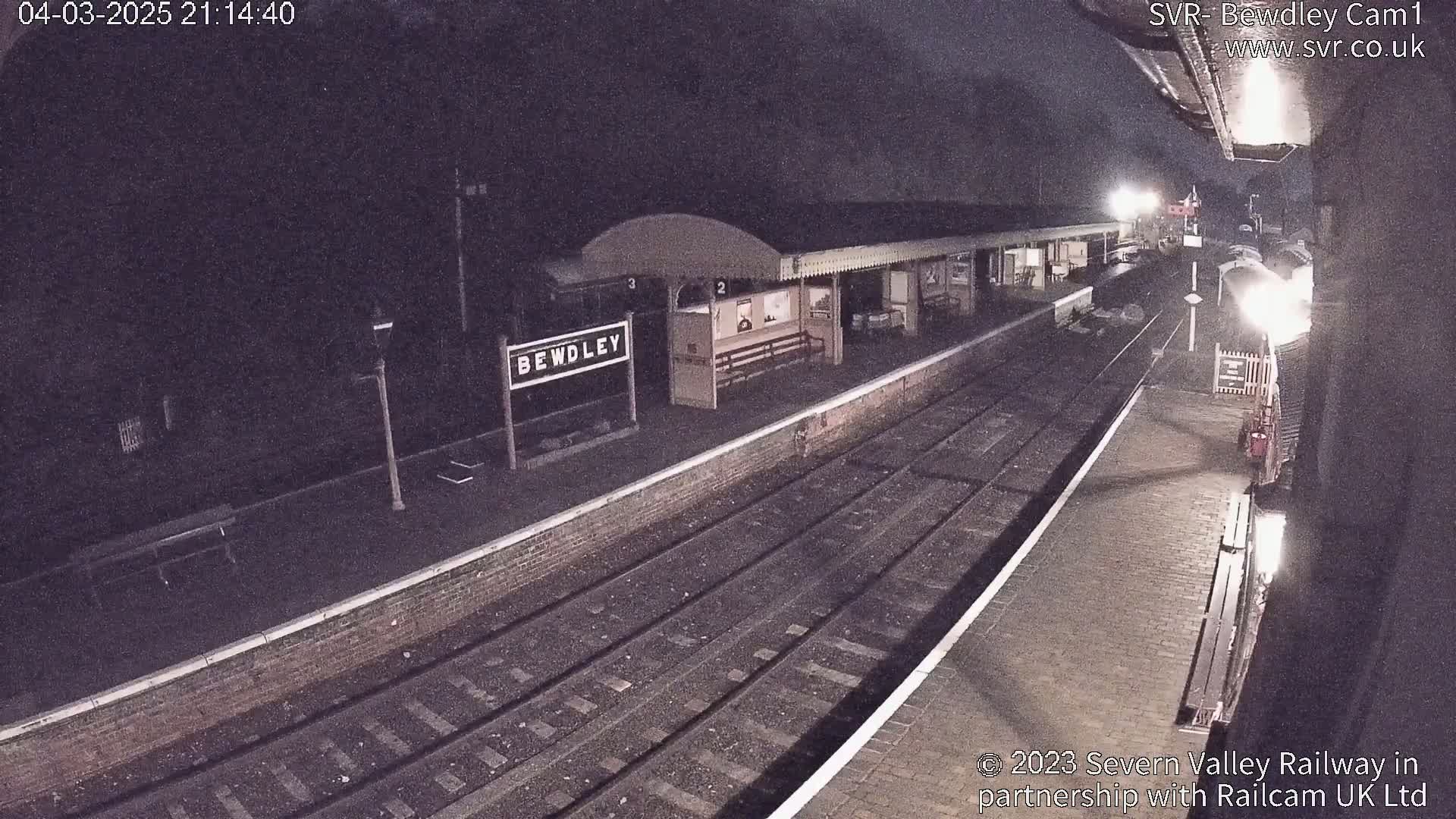 A nighttime view of a train station platform with a train partially visible in the distance under dark conditions.