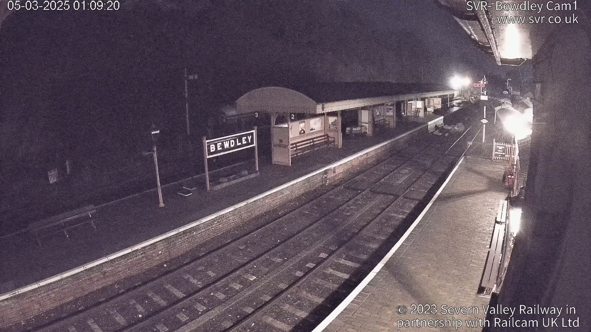 A nighttime view of a train station platform with train tracks and a partially visible train.
