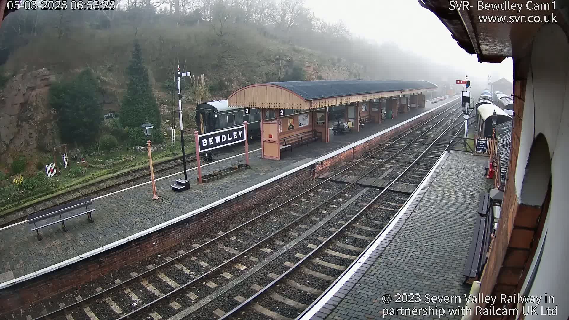A train station platform on a foggy morning with a train partially visible and a covered waiting area.