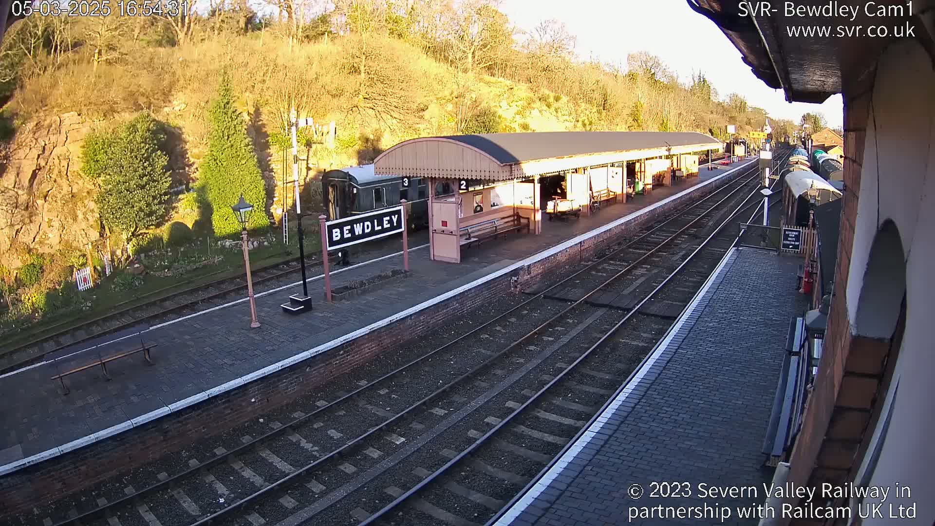 A train sits at a station platform on a sunny day, next to a brick building and a rock face.