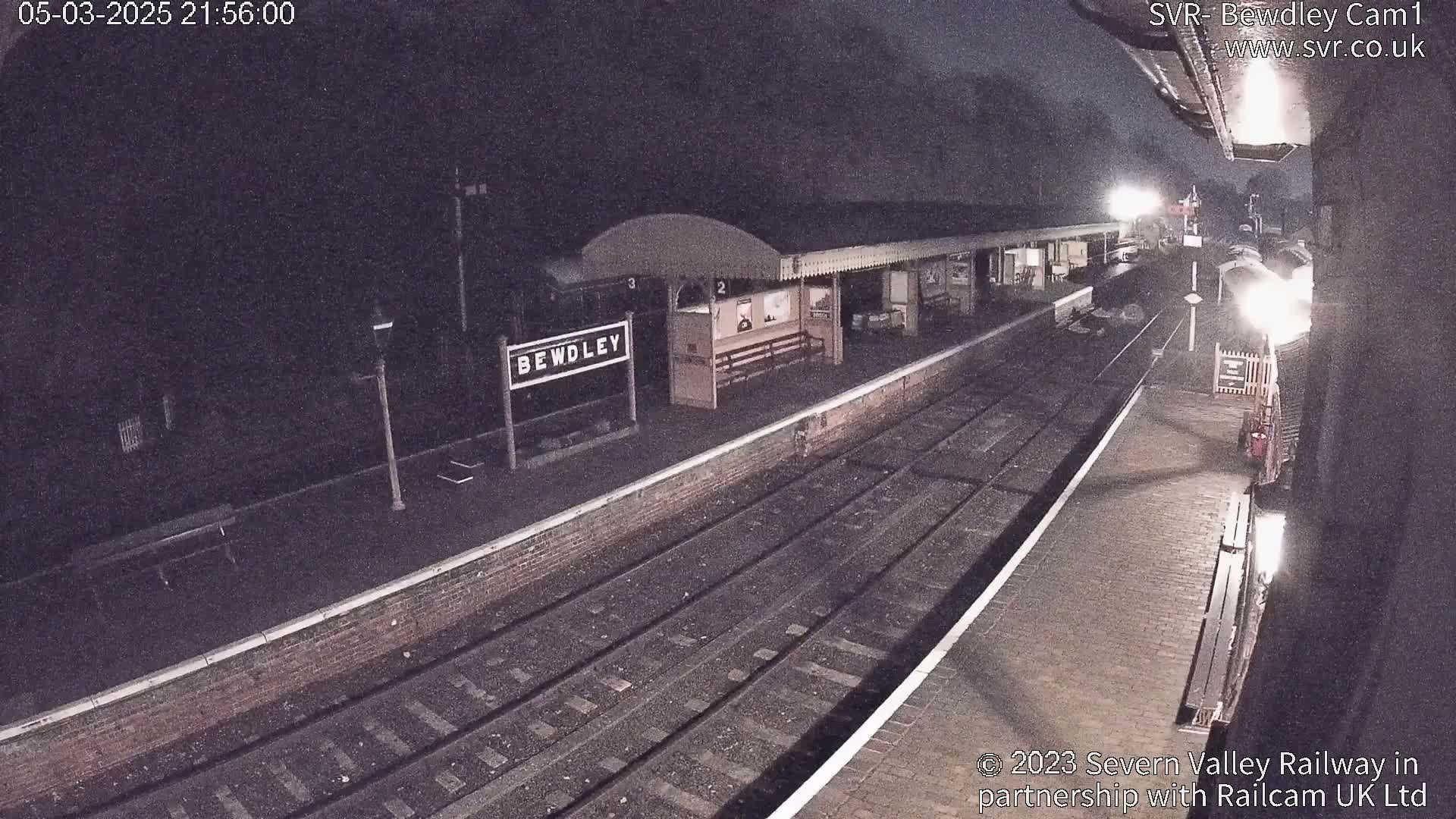 A nighttime view of a train station platform with train tracks and a partially visible train, under dark conditions.