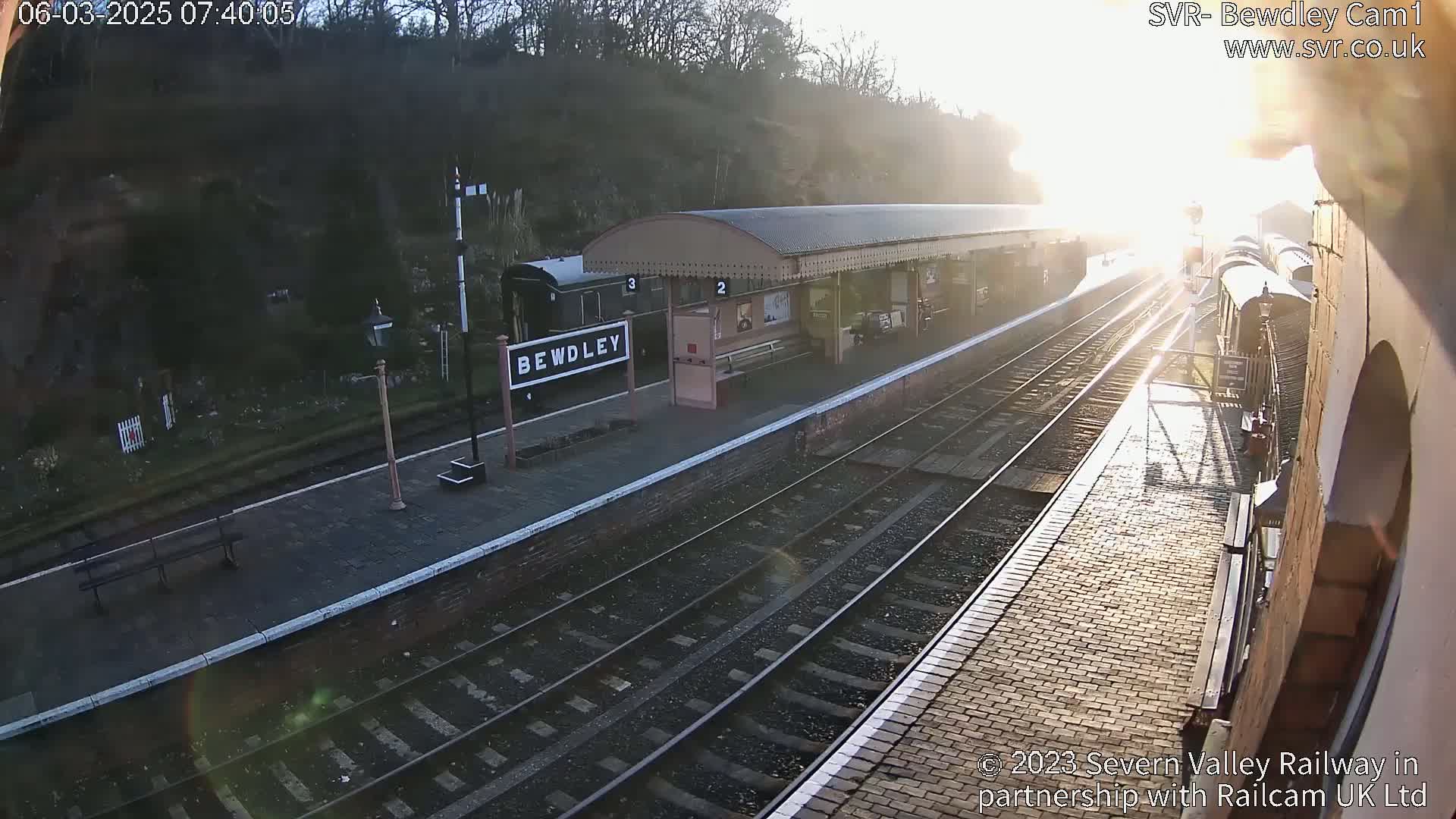 A train station platform with a train arriving in the sunlight, on a sunny morning.