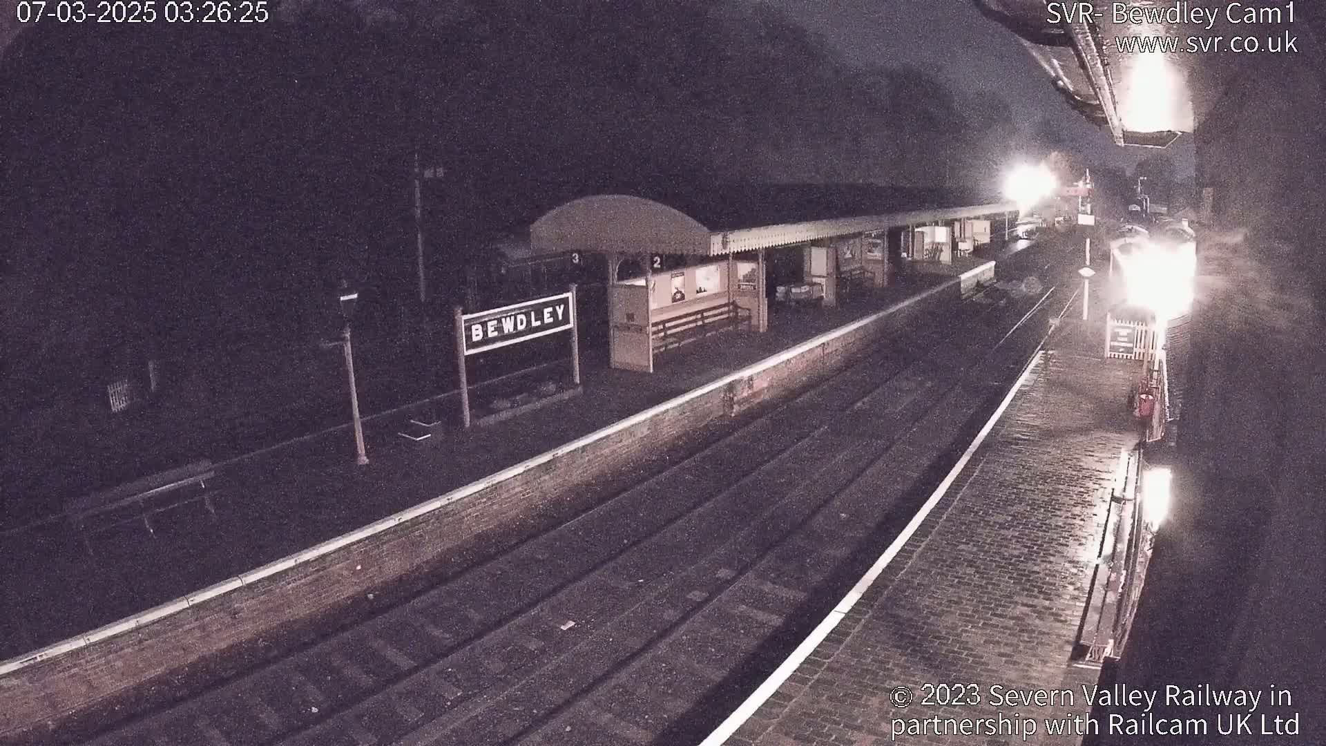 A train sits at a dimly lit, rain-slicked nighttime train station.