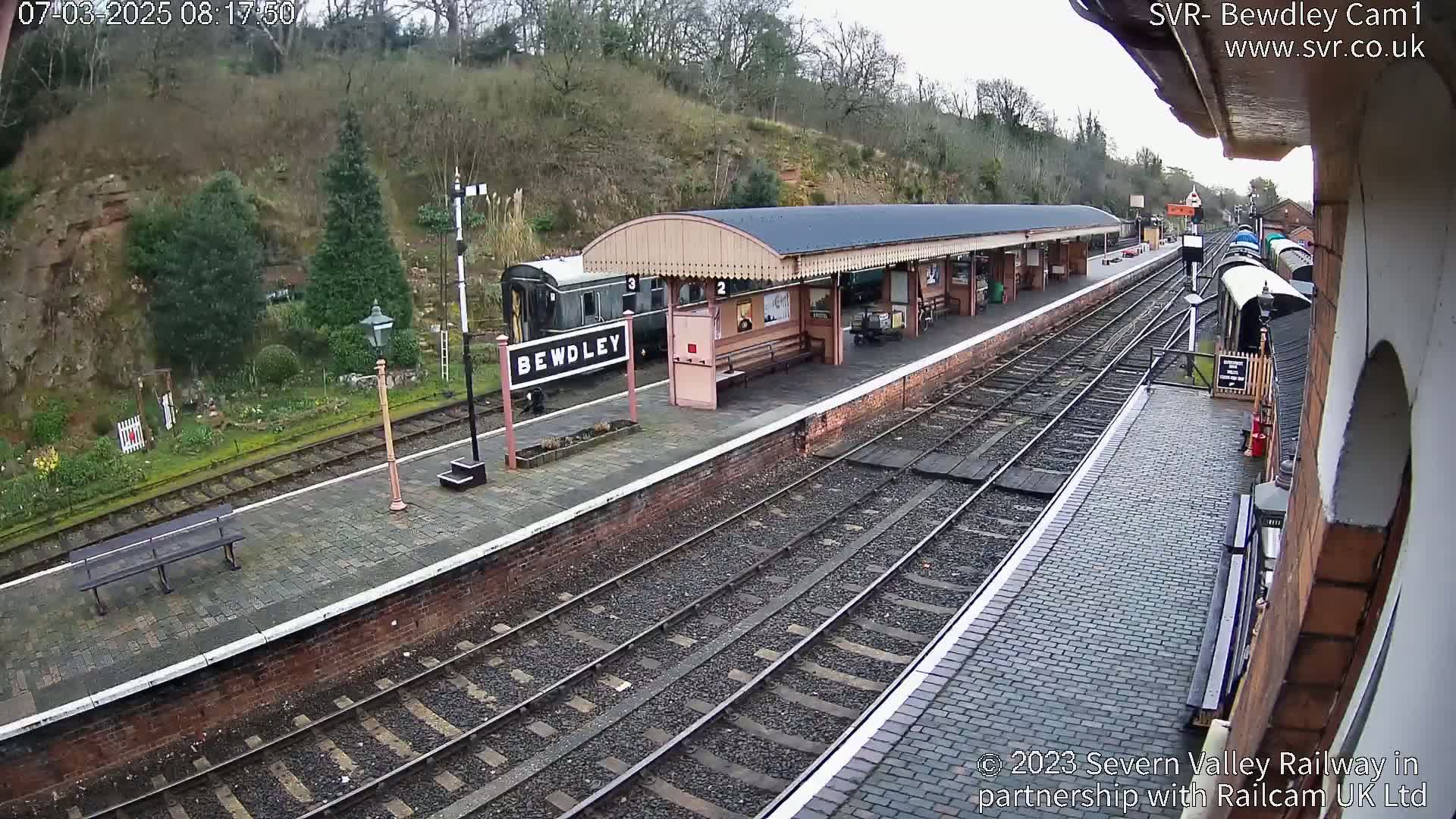 A train station platform with a railway line, train cars, and a covered waiting area on an overcast day.