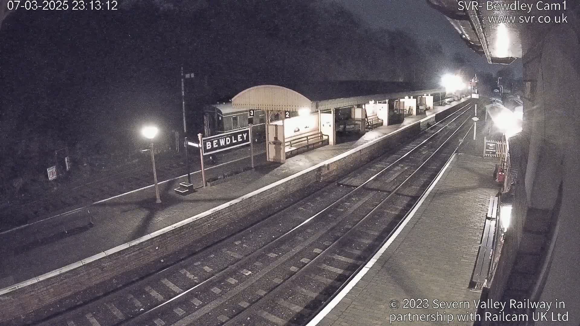 A nighttime view of a train station platform with a train car visible, under light rain or snowfall.