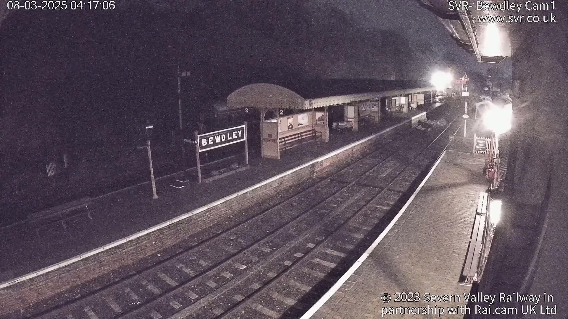 A nighttime view of a train station platform with train tracks and a partially visible train under a dark, possibly cloudy sky.
