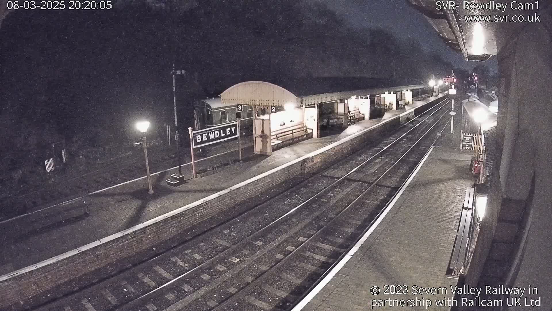 A nighttime view of a train station platform with a train partially visible, lit by several lamps under a dark sky.