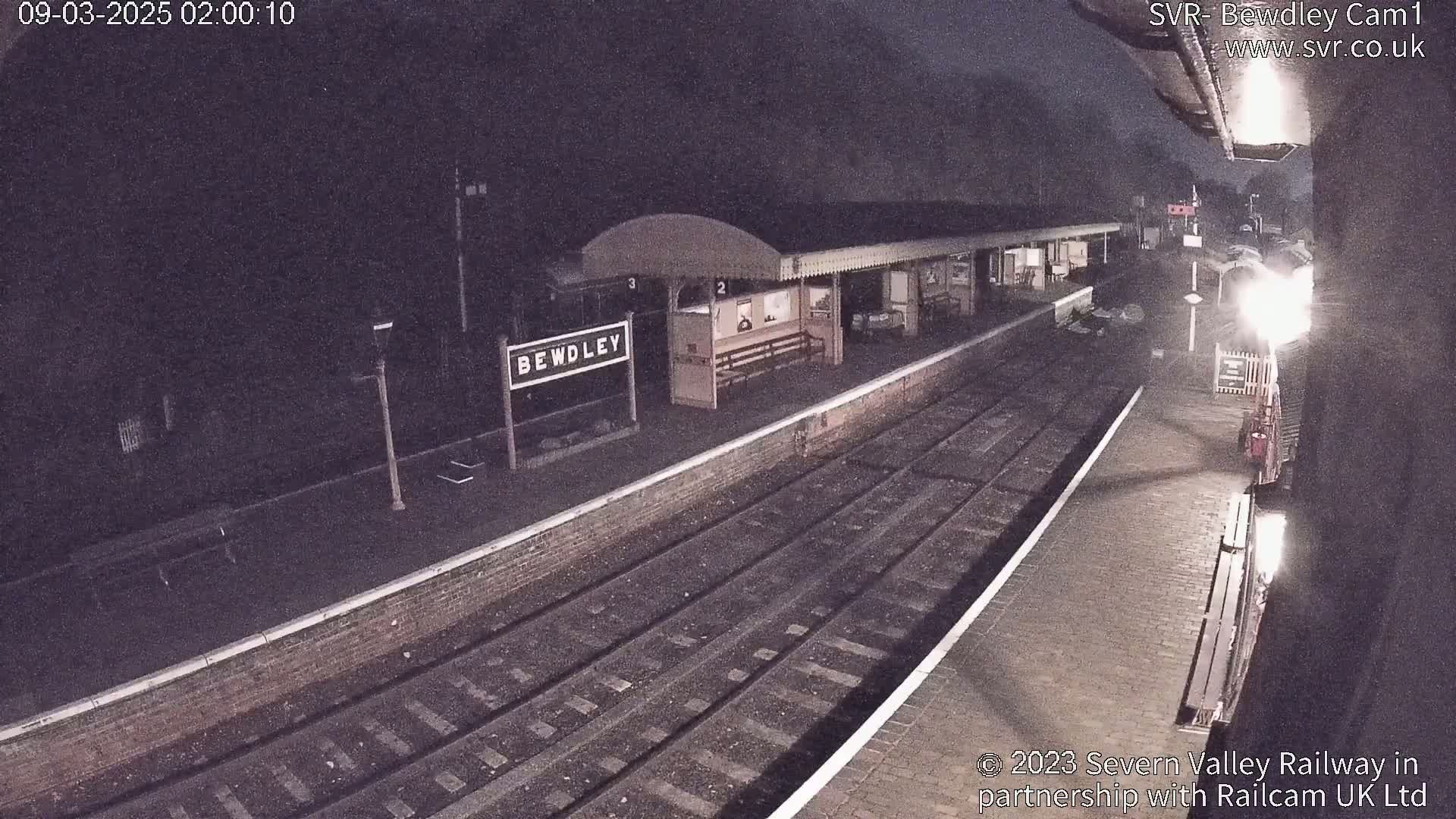 A nighttime view of a train station platform with train tracks and a partially visible train under dim lighting.