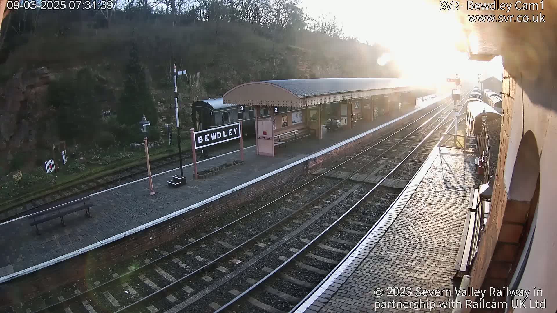 A sunny morning at a train station with railway tracks and passenger cars visible, showing a brick platform and a covered waiting area.