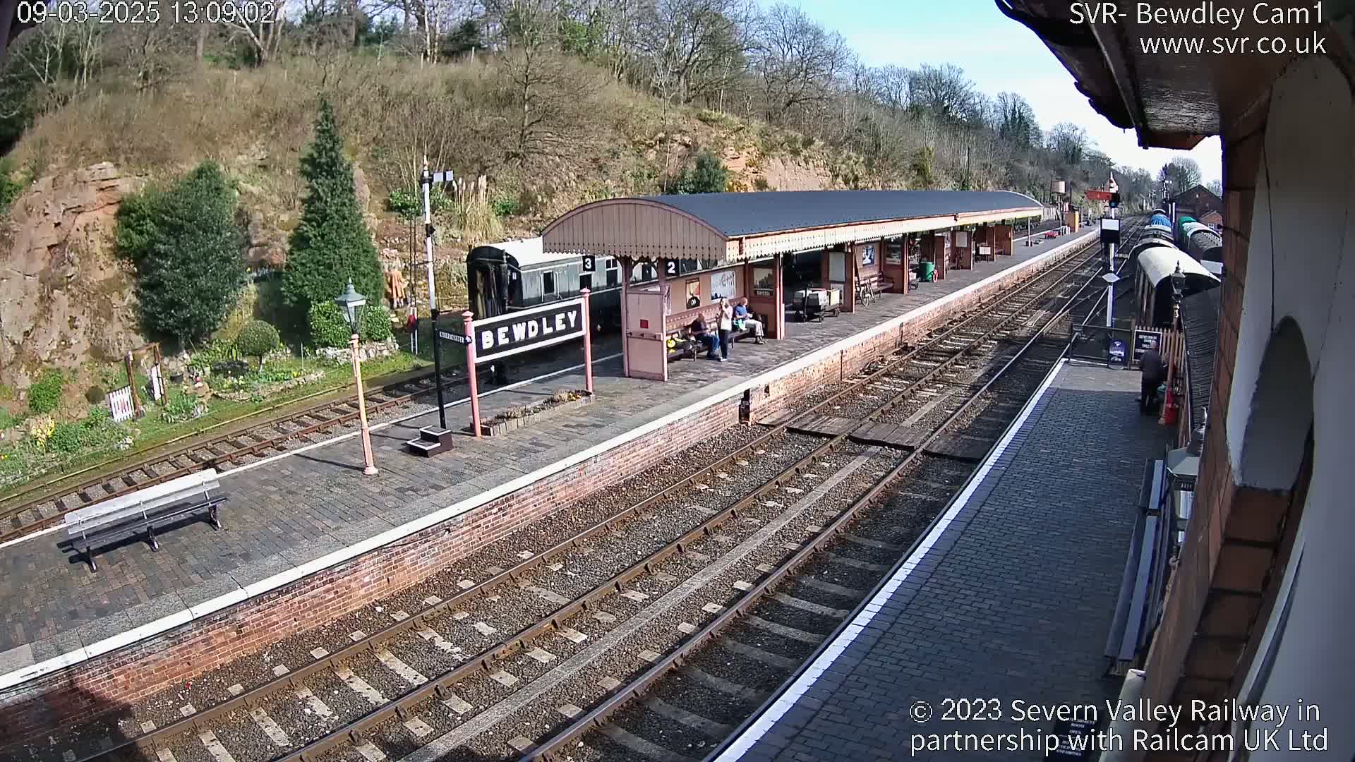 A train station platform with a train and several people waiting under a covered shelter on a sunny day.