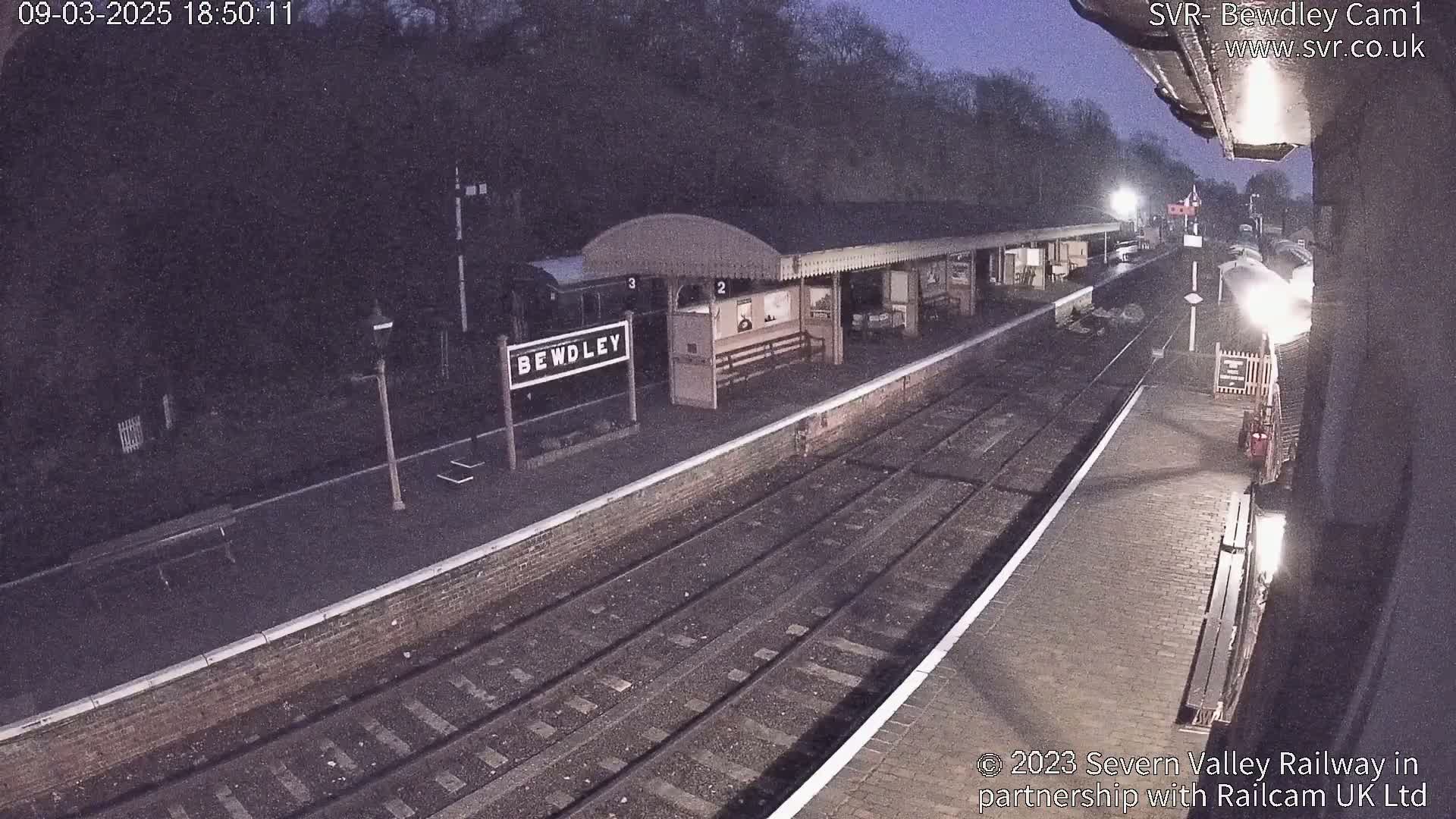 A dimly lit train station at night, with train tracks and a partially visible train, under a dark sky.