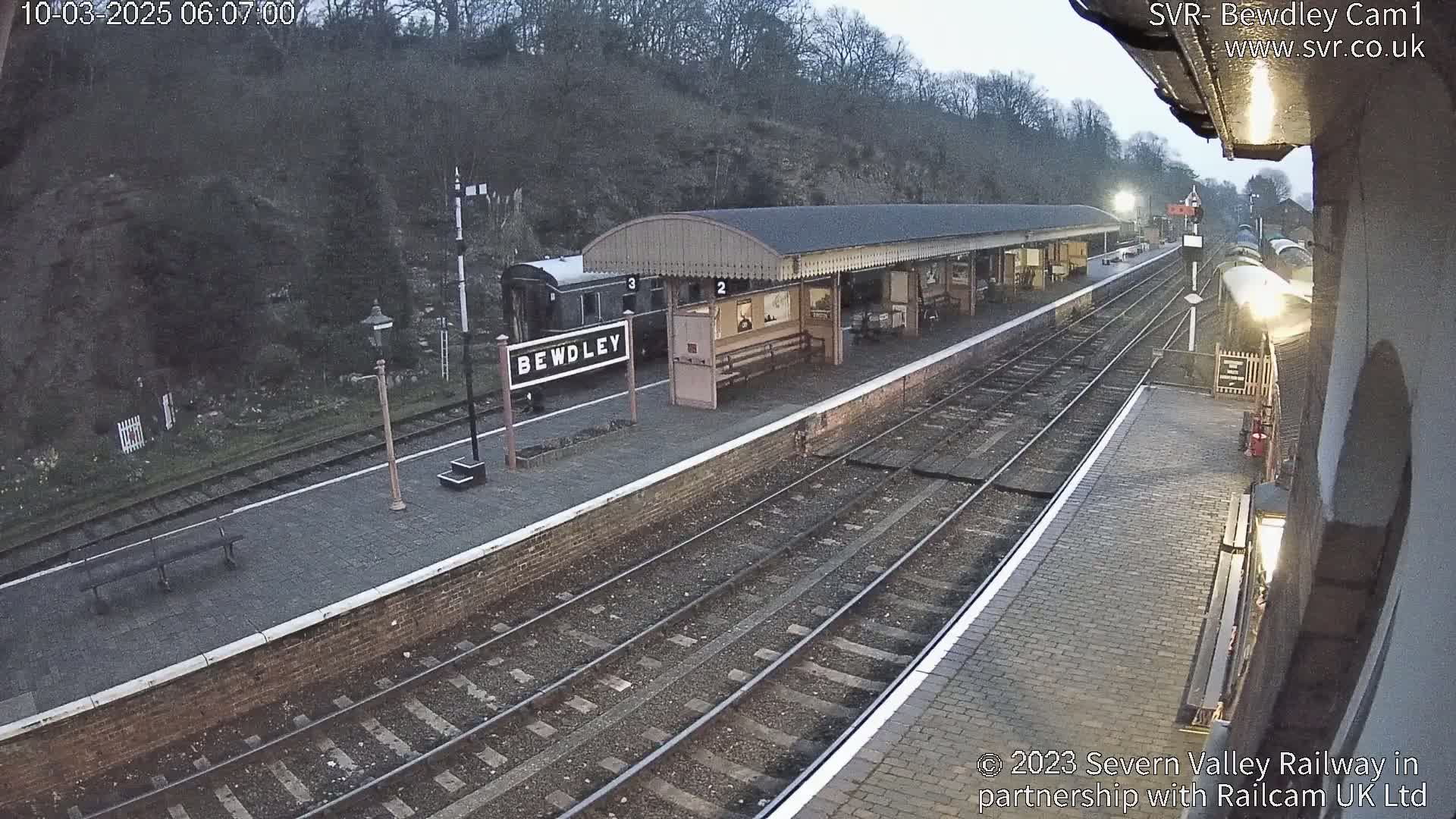 A train station platform with a covered waiting area, train tracks, and a train partially visible, under overcast conditions.