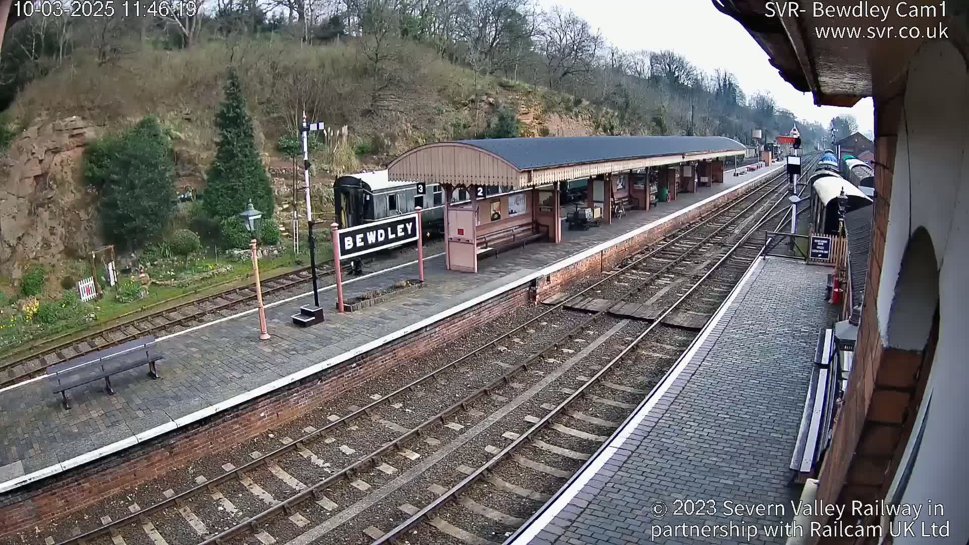 A train sits at a railway station platform next to a covered waiting area, with a hillside visible beyond the tracks in an overcast outdoor setting.