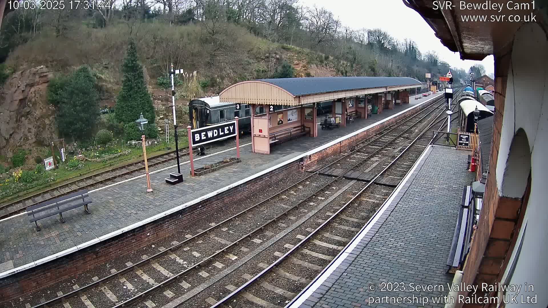 A train sits at a station platform next to a covered waiting area on an overcast day.