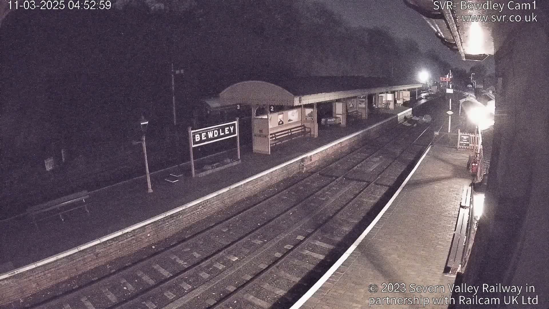 A nighttime view of a train station platform with train tracks and a partially visible train under dark, nighttime conditions.