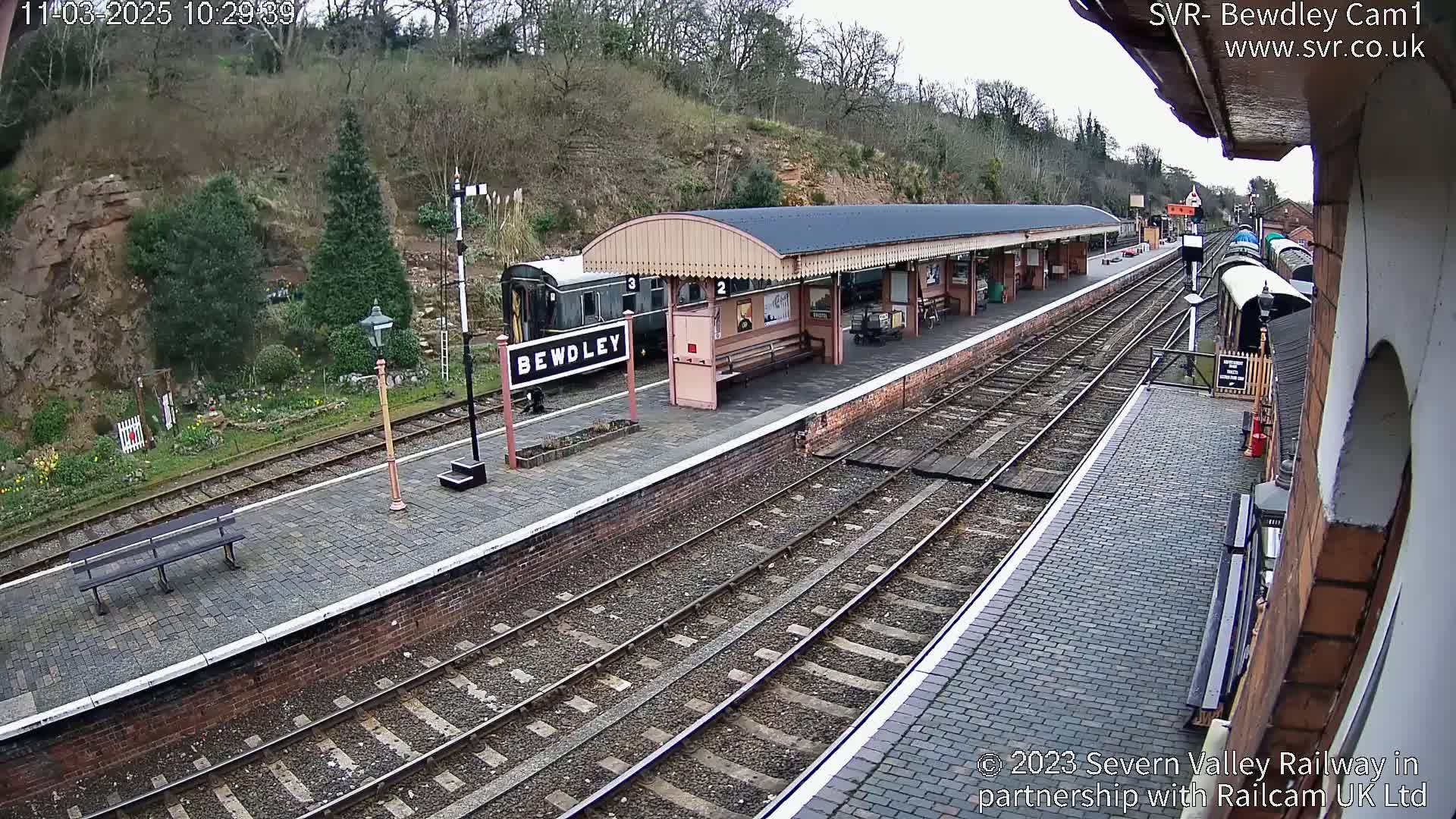 A train station platform with a covered waiting area, railway tracks, and a partially visible train, all under an overcast sky.