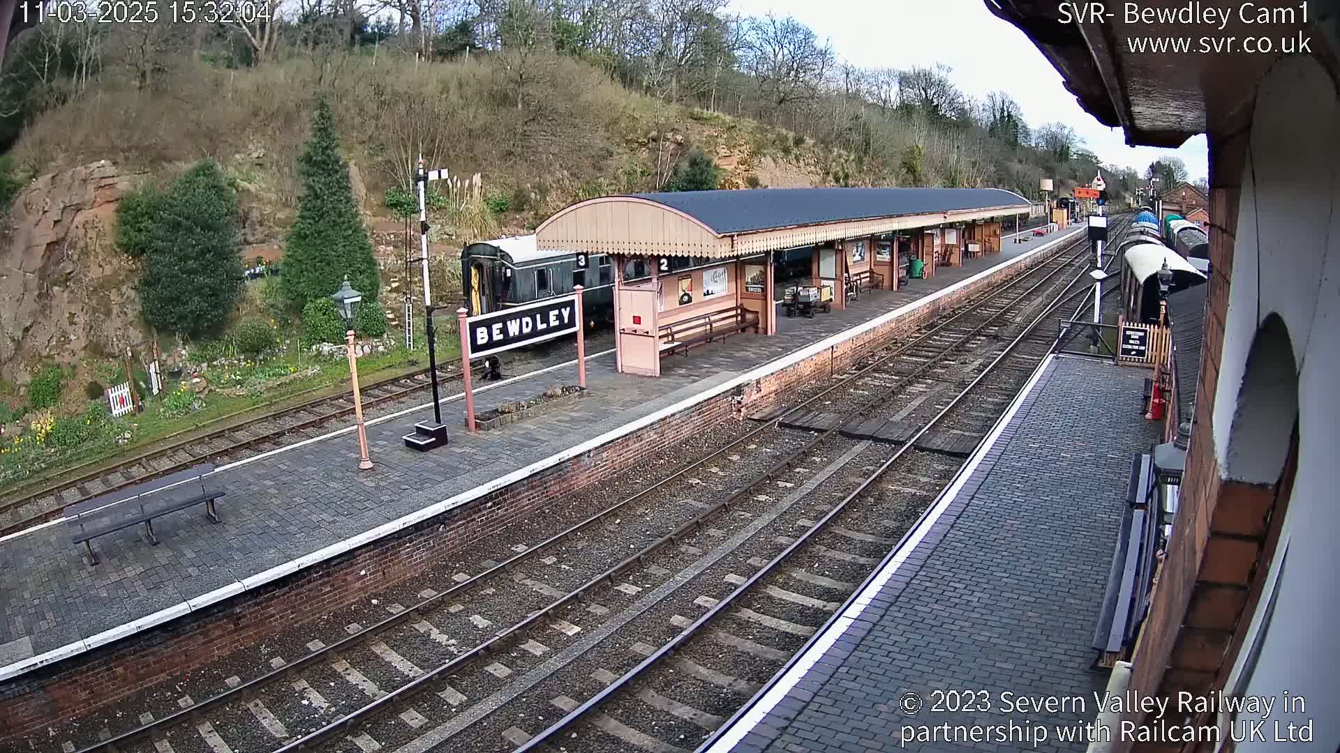 A train sits at a railway station platform on a partly cloudy day.