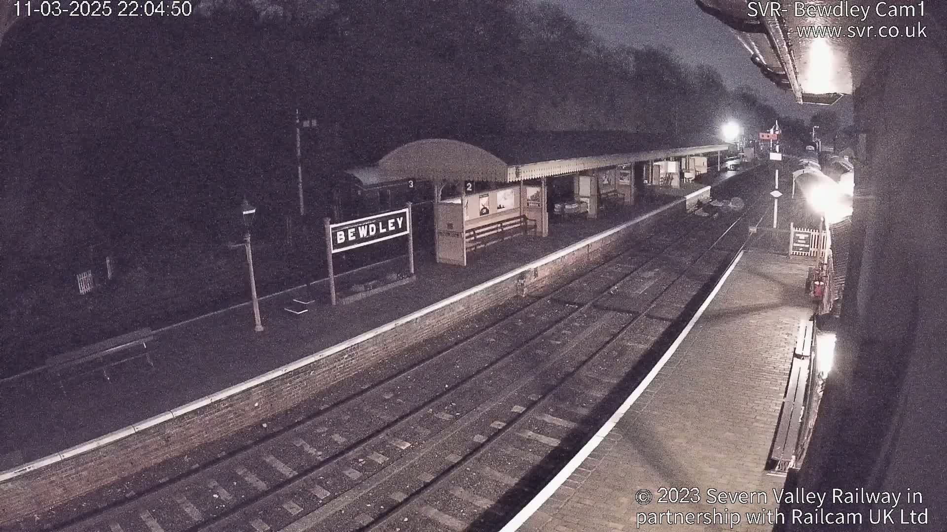 A nighttime view of a train station platform with train tracks and a partially visible train under a dark, possibly cloudy sky.