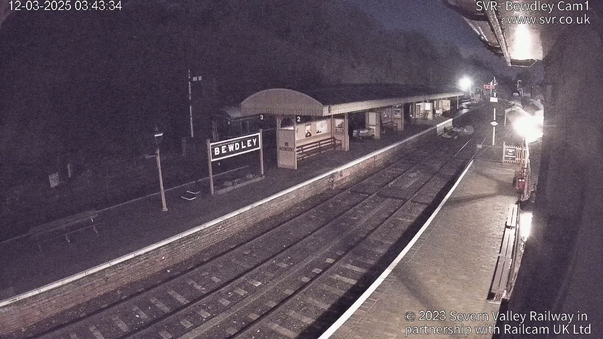 A nighttime view of a train station platform with train tracks extending into the darkness.