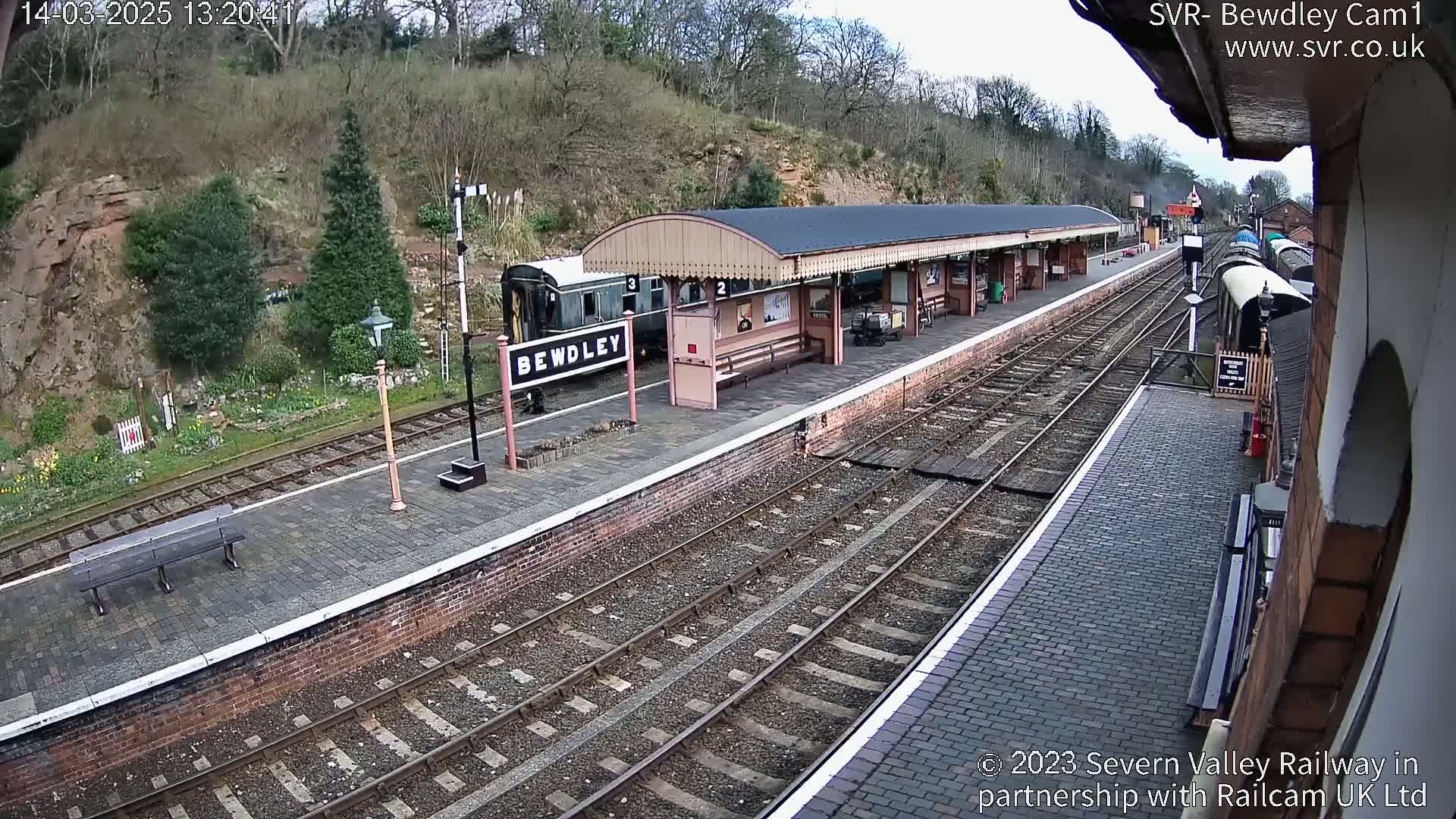 A railway station platform with a train and covered waiting area, on a partly sunny day.