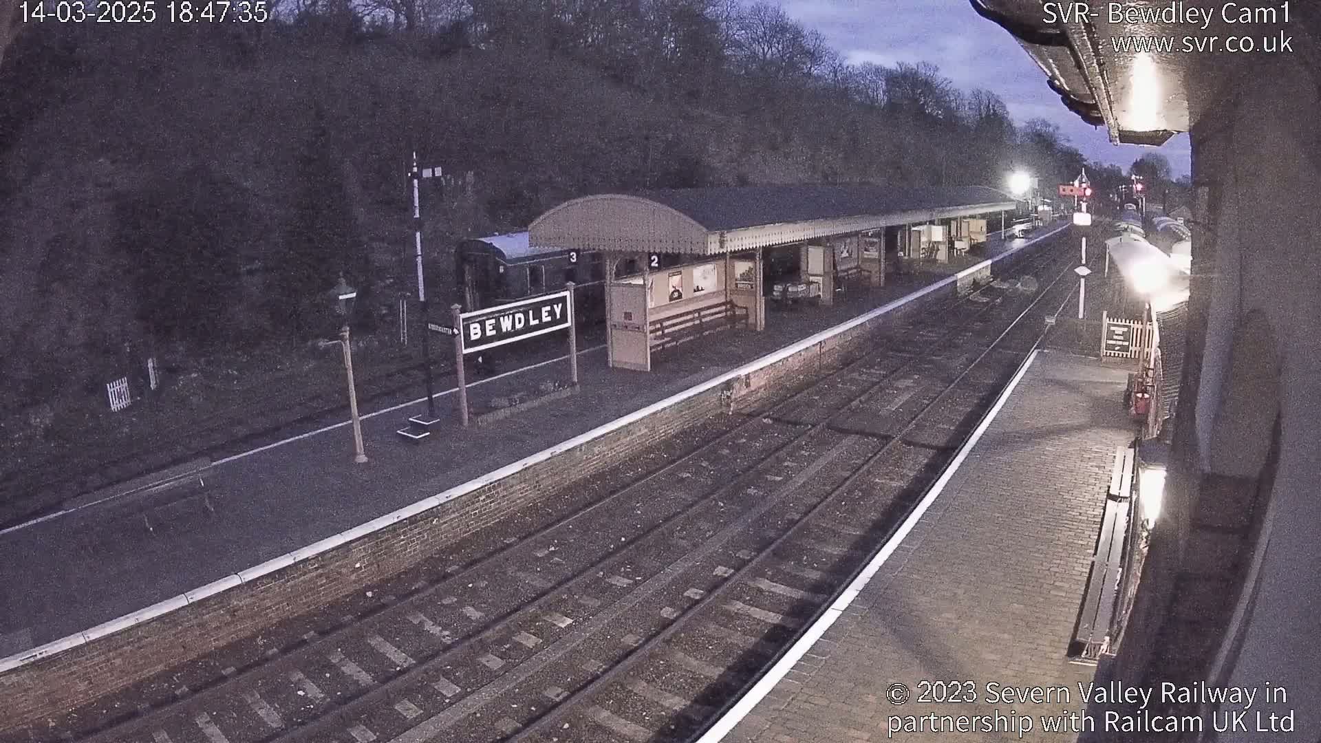 A nighttime view of a train station platform with a train partially visible and a covered waiting area, under a dark sky.
