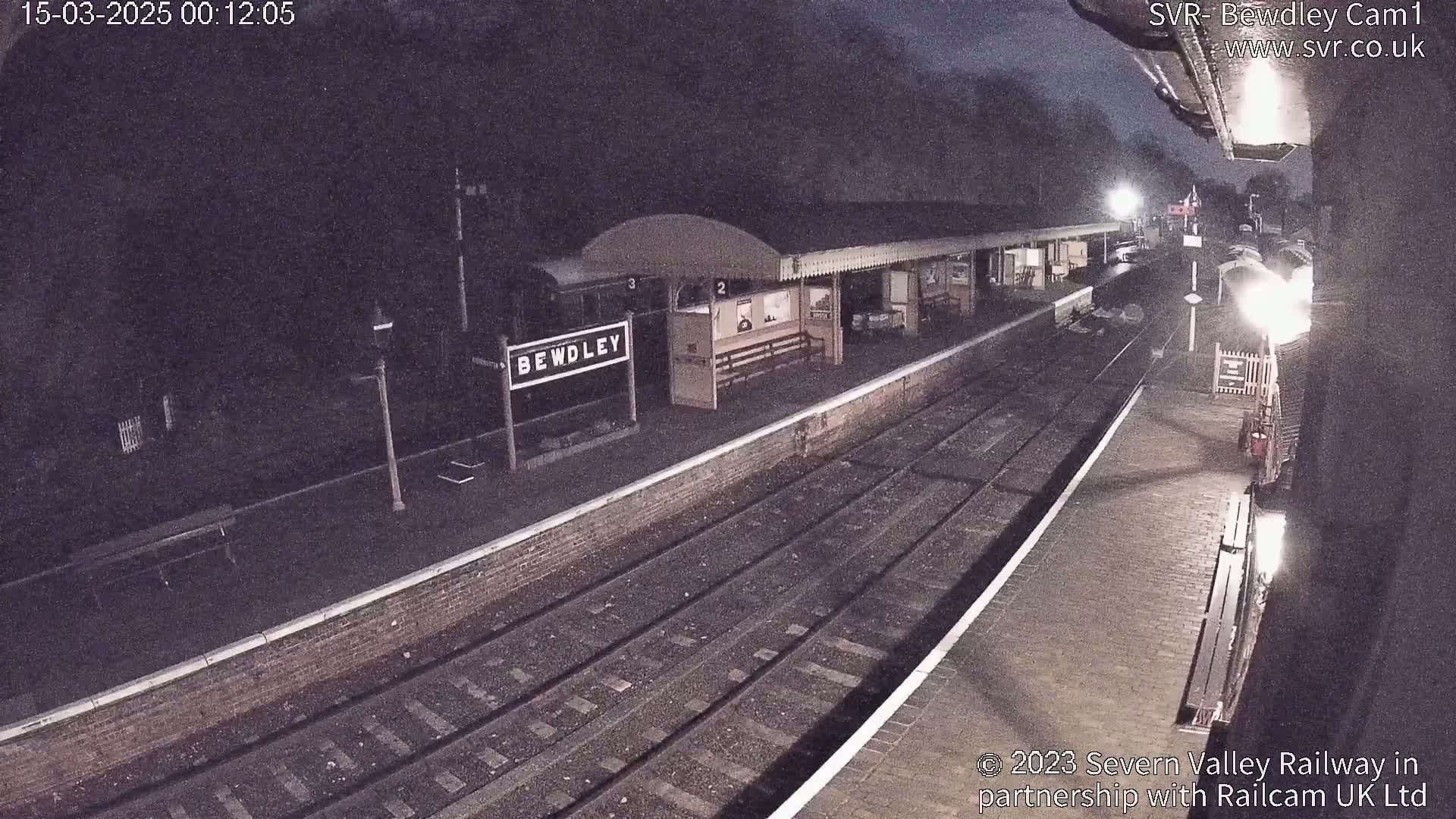 A nighttime view of a train station platform with train tracks, a shelter, and a sign indicating "Bewdley."
