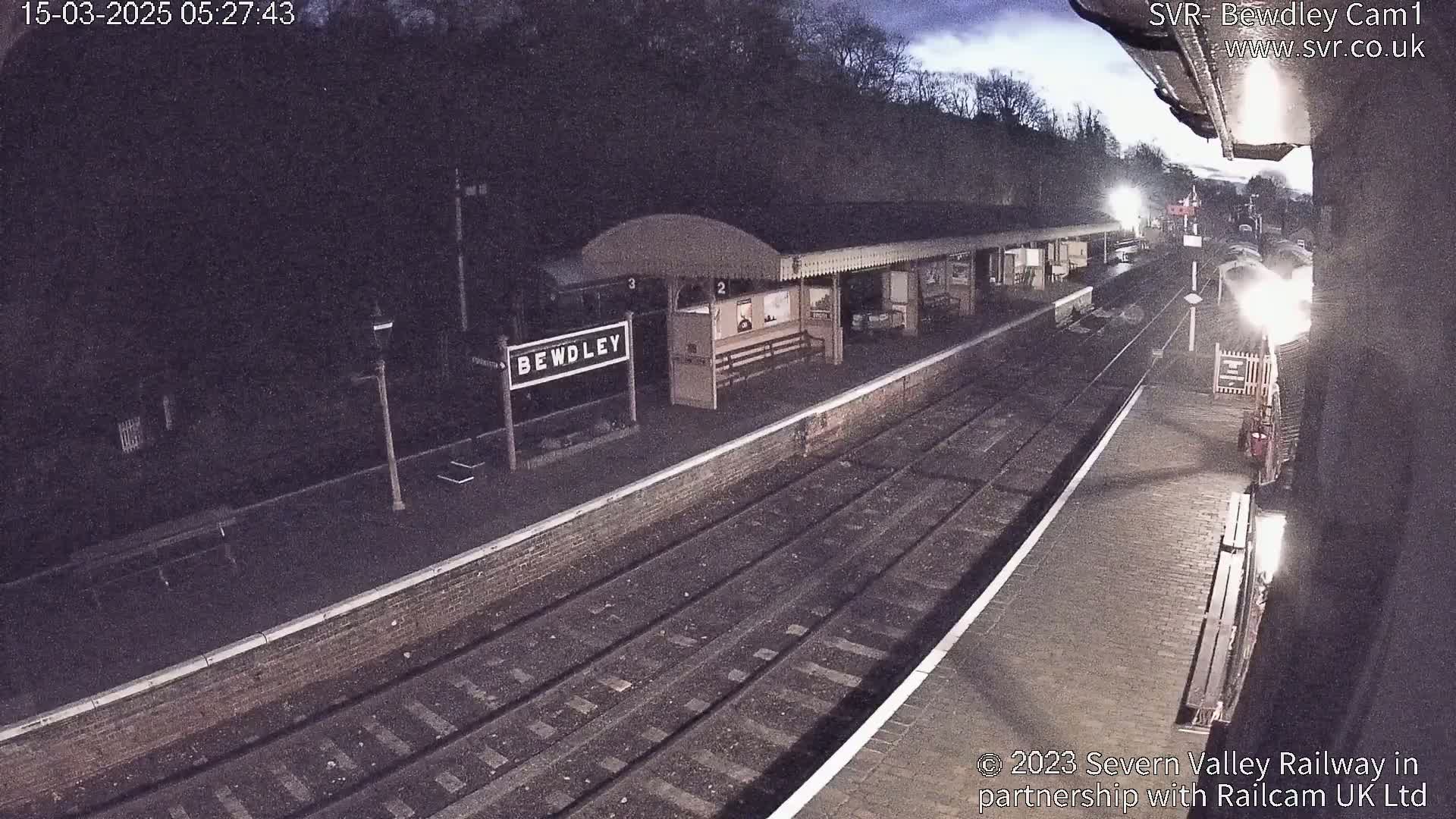 A nighttime view of a train station platform with multiple train tracks, a station building, and a partially visible train in the distance under a dark, cloudy sky.