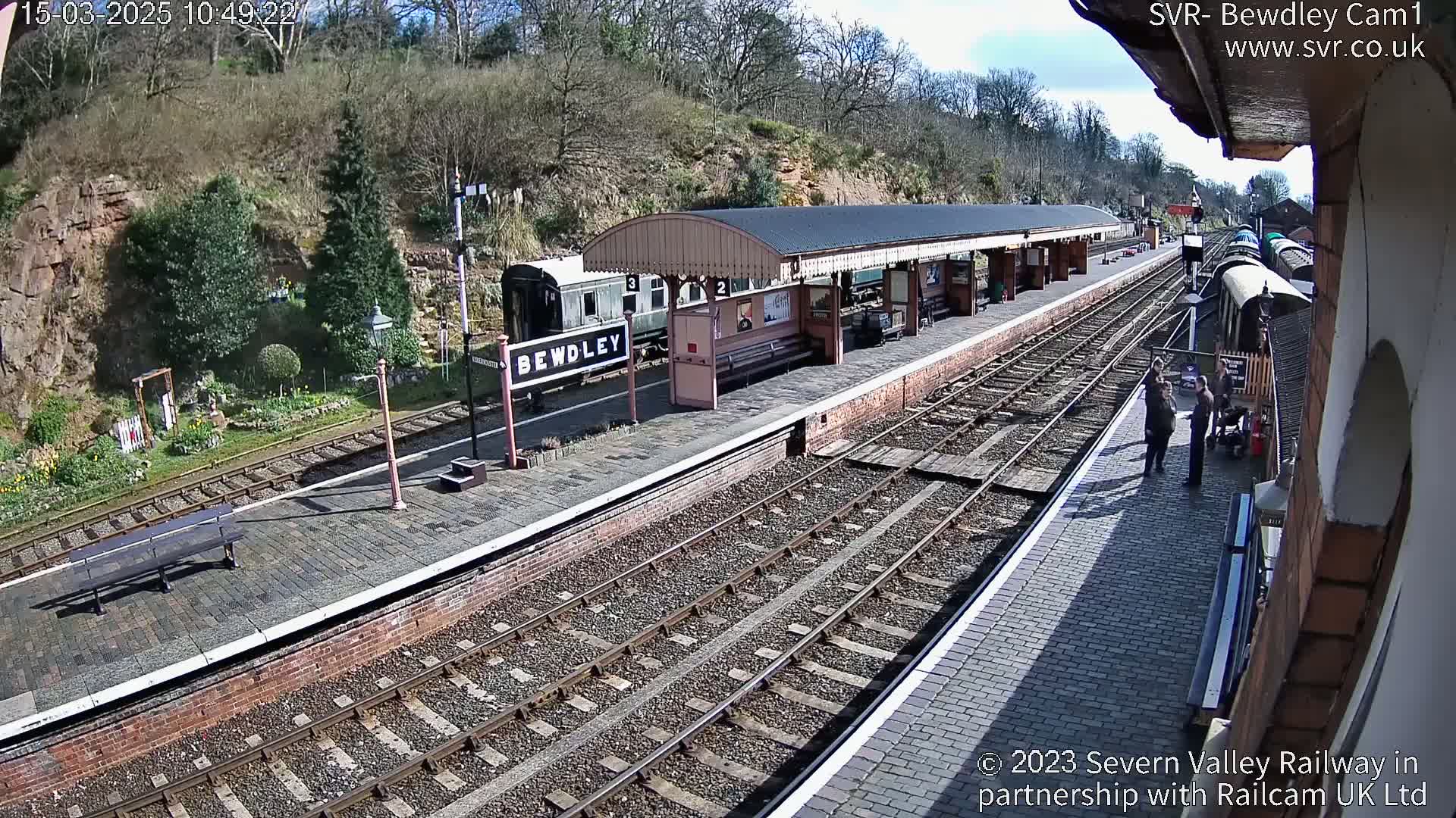 A sunny day at a train station with multiple train tracks, a platform with a covered waiting area, and several people standing near a railway carriage.