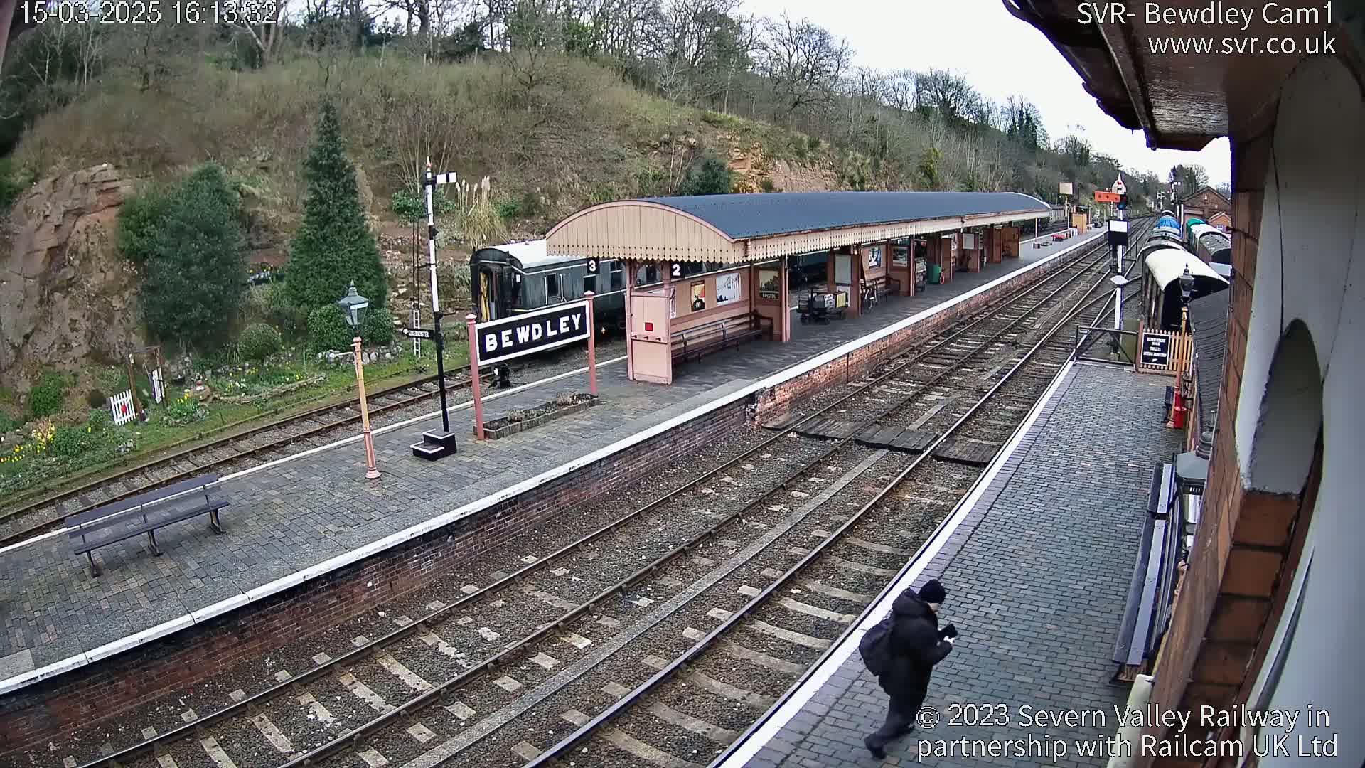 A train station with a covered platform, a train car, and a person walking on a paved path under a cloudy sky.