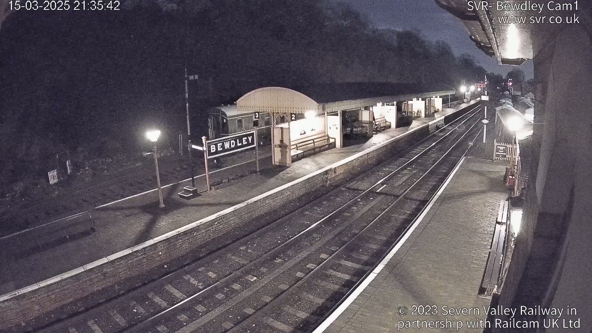 A nighttime view of a train station platform with train cars, tracks, and a lit lamppost.