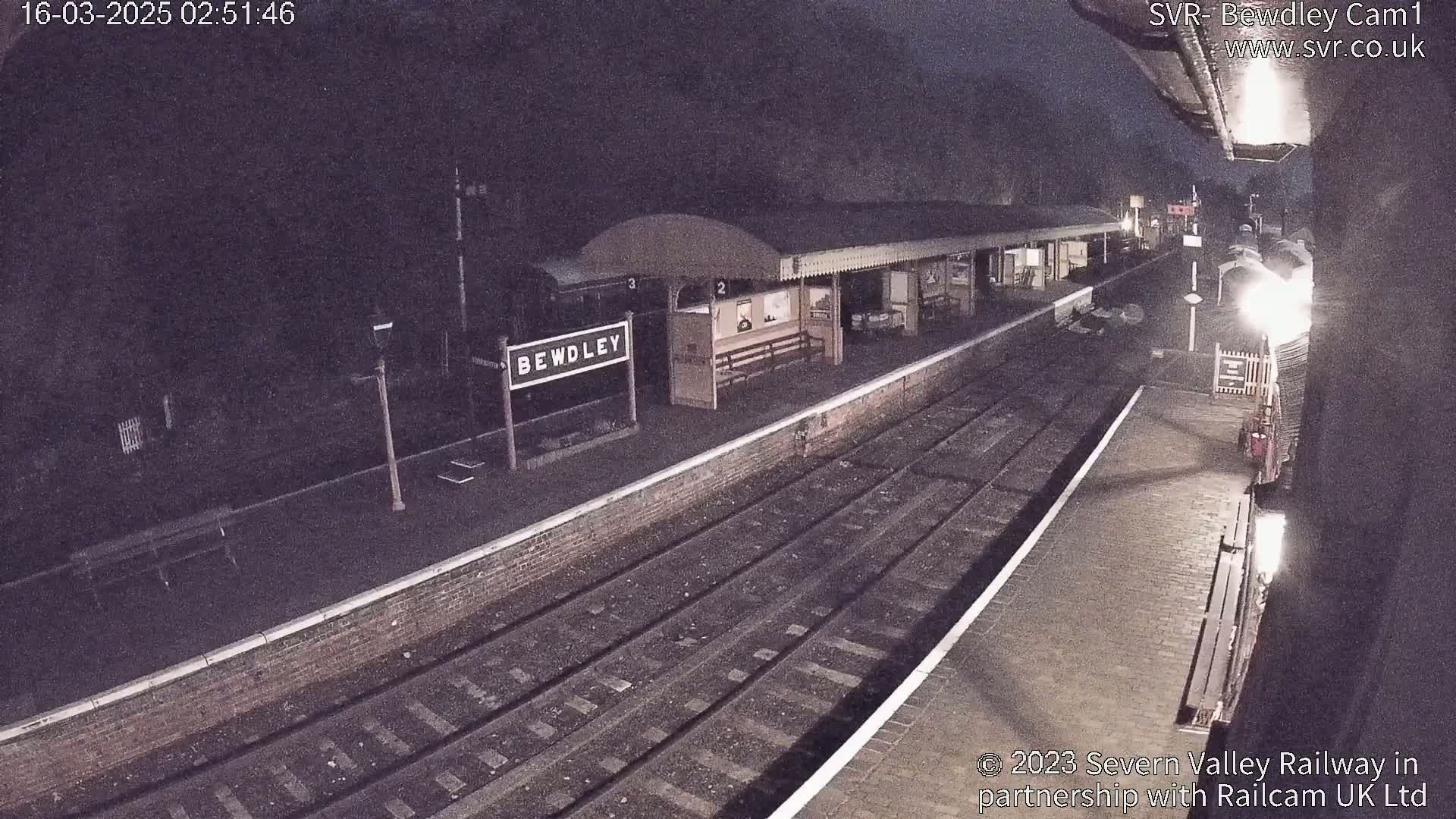 A nighttime view of a train station platform with a train partially visible, under dark skies.