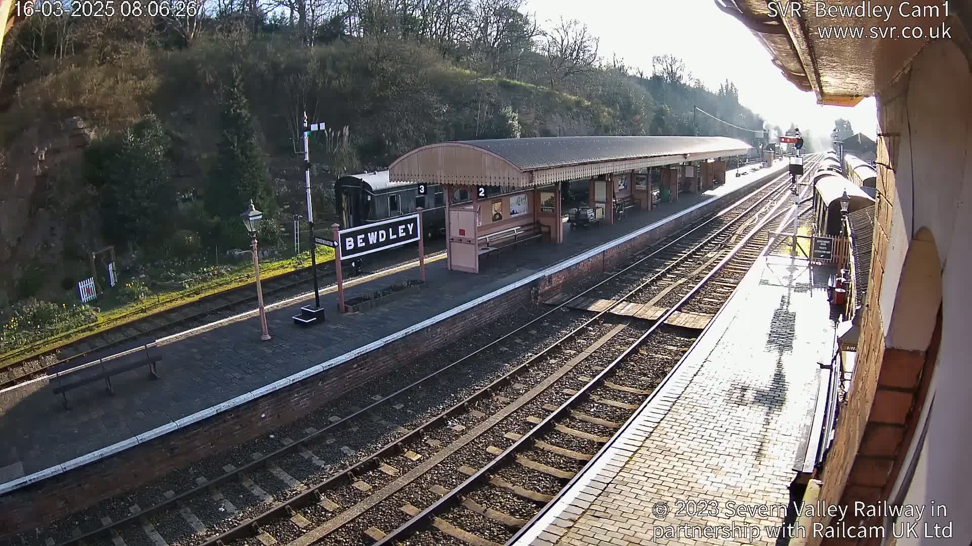 A sunny view of a train station platform with railway tracks, a train, and a covered waiting area.