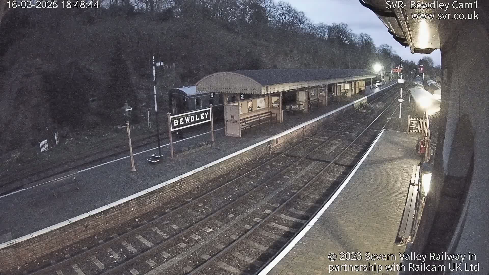 A train sits at a dimly lit, brick railway station platform under a twilight sky.