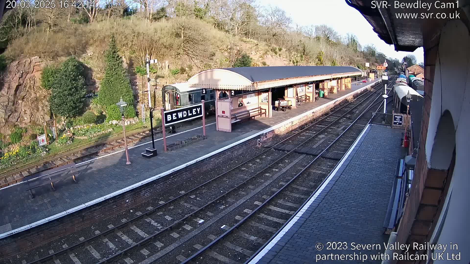 A train sits at a station platform next to a covered waiting area on a sunny day.