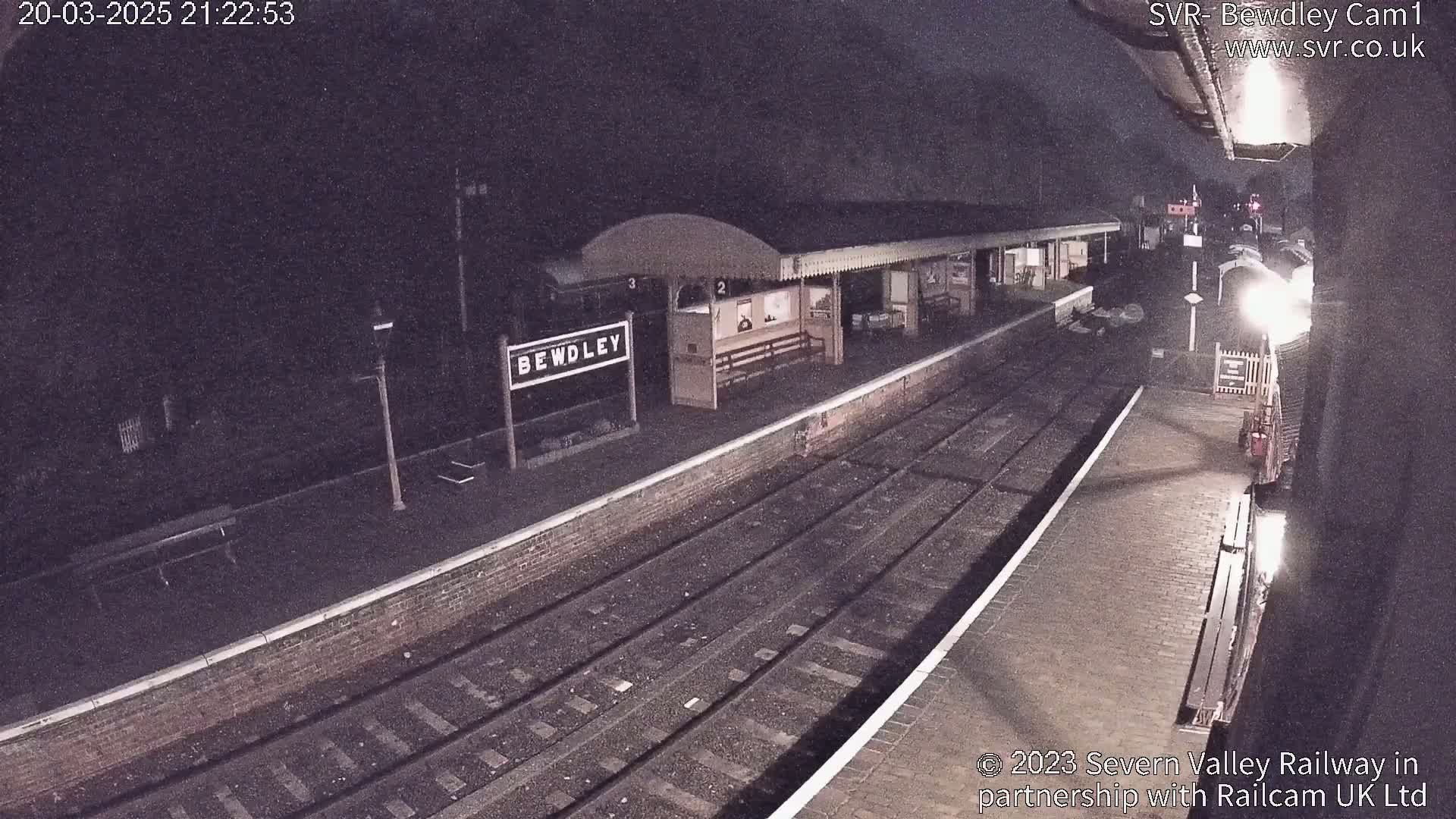 A mostly empty, nighttime train station platform with train tracks and a partially visible train.
