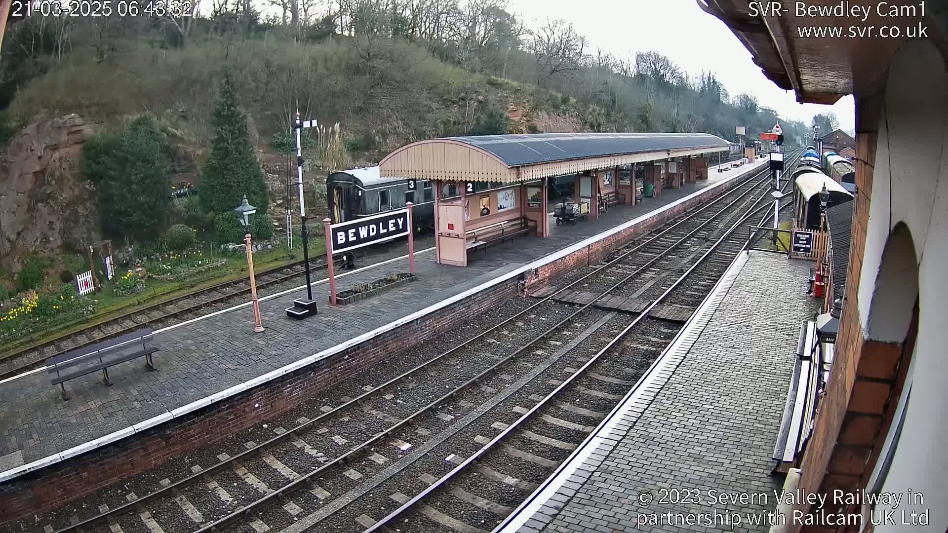 A train station platform with several railway tracks, a partially visible train, and a covered waiting area is seen on an overcast day.