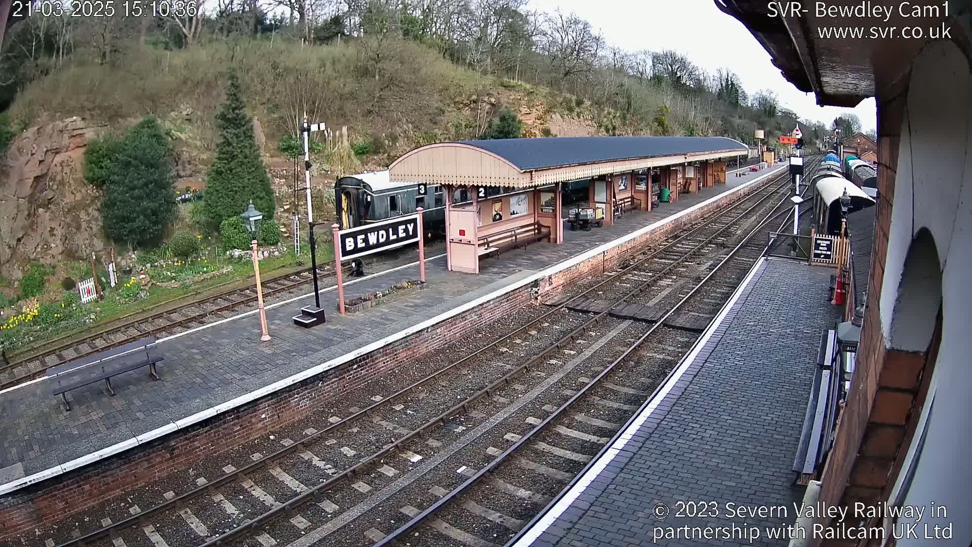 A train is stopped at a railway station platform under a covered awning on a partly sunny day.