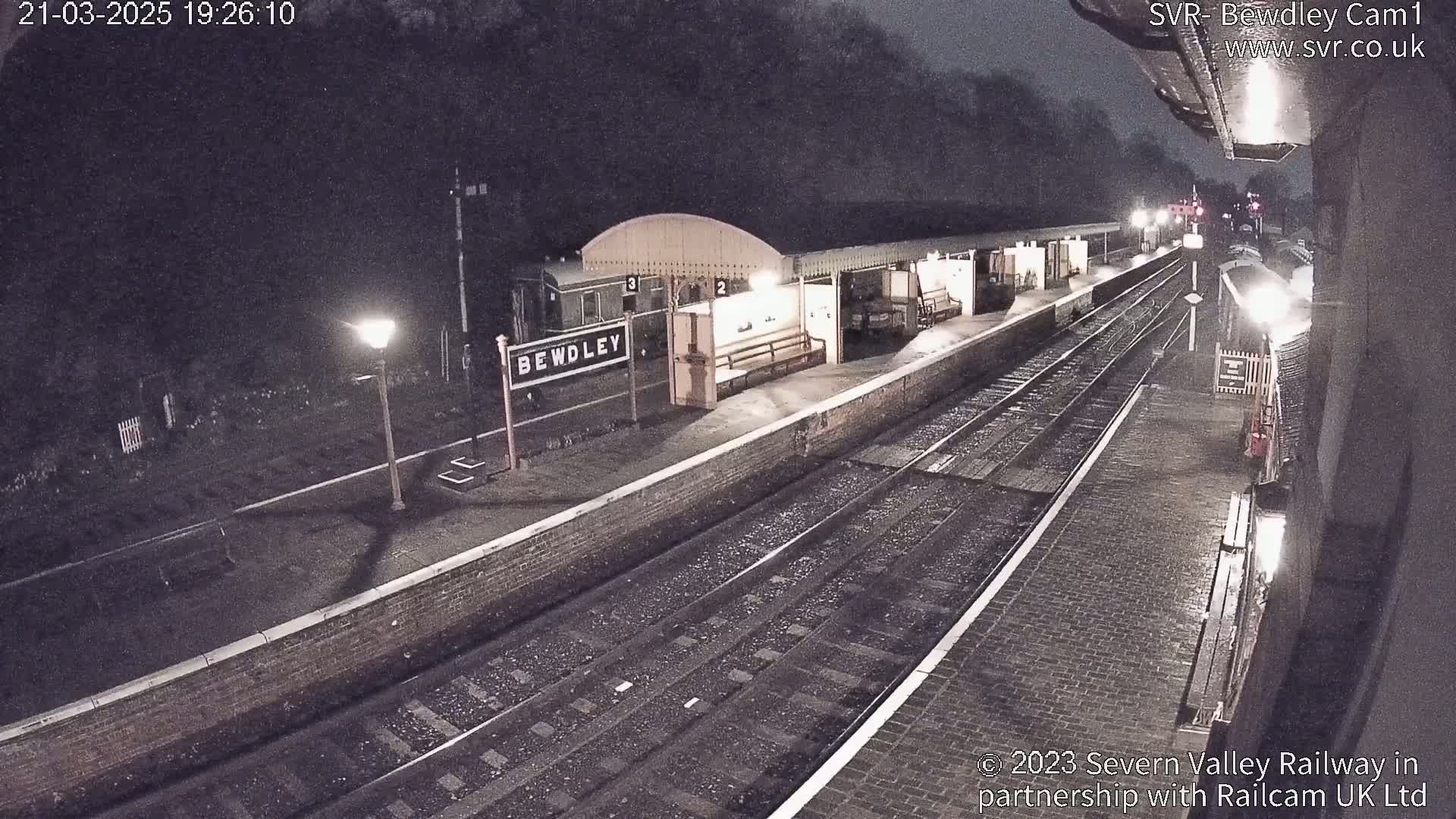 A nighttime view of a train station platform with train tracks, platforms, and lights, under light snowfall.